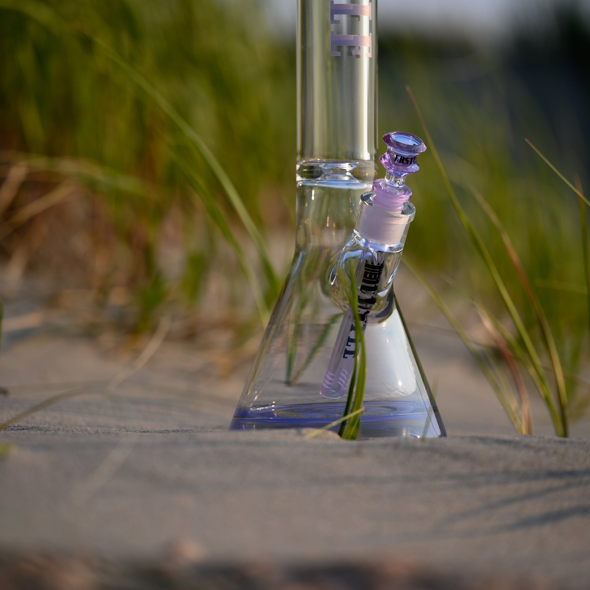 A clear glass beaker bong with purple accents, featuring the "Castle" logo vertically on the neck. The bong is set on sandy ground with green grass blades surrounding it, creating a natural beach-like setting. The sunlight highlights the clarity and craftsmanship of the glass, casting soft shadows and enhancing the overall aesthetic appeal. The scene blends the sleek design of the bong with the serene and earthy elements of nature.