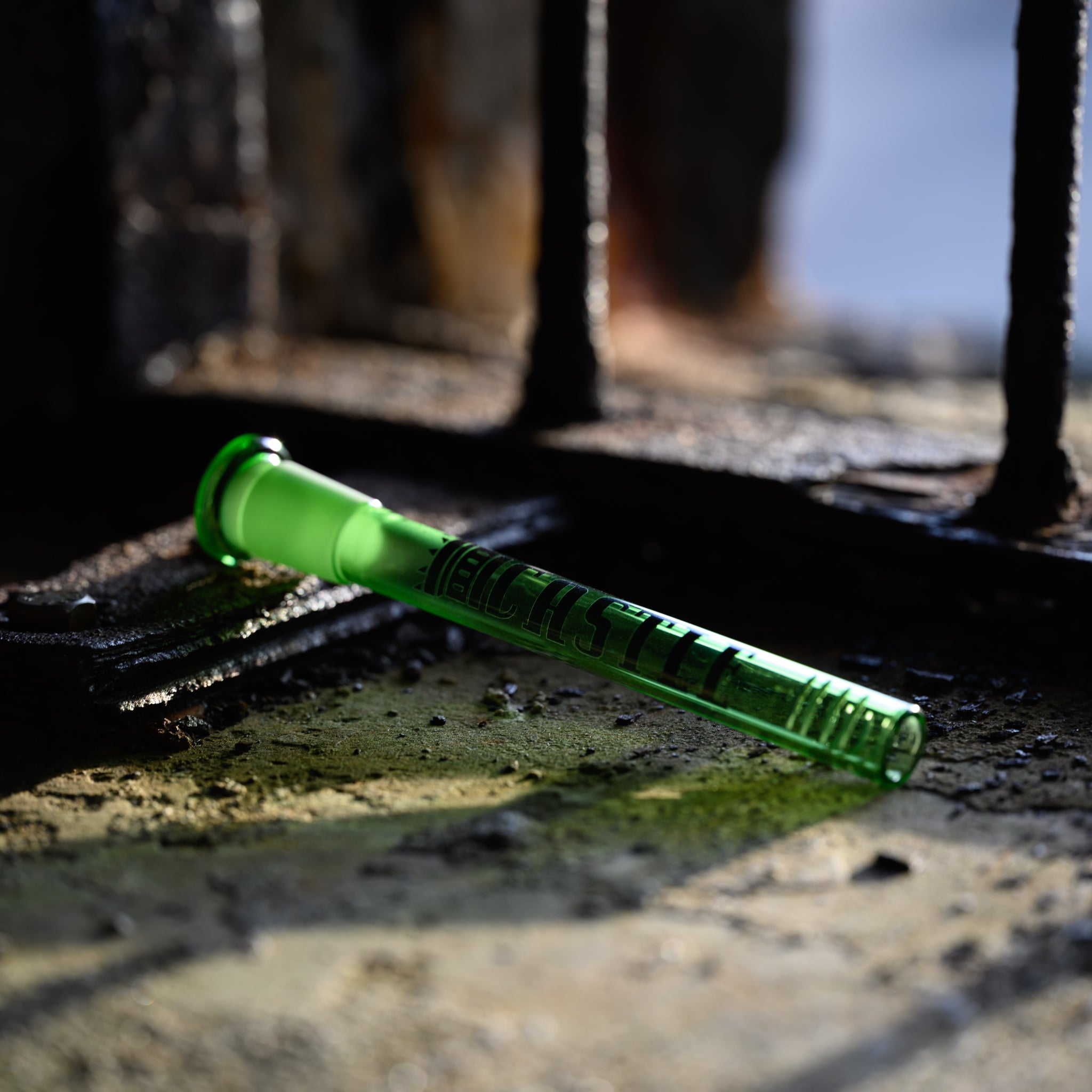 A green glass downstem with the Castle Glassworks logo lies horizontally on a gritty, industrial surface, illuminated by soft natural light streaming through iron bars in the background. The downstem features intricate details and 8 slits, casting subtle green reflections on the surrounding area.