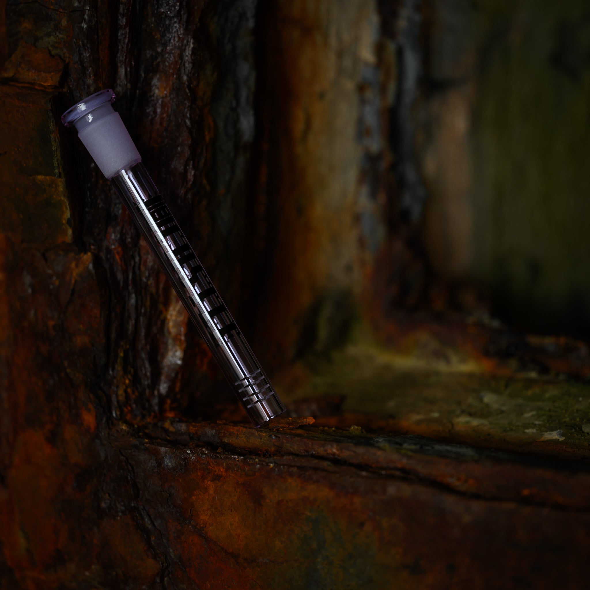 A sleek, purple glass downstem with the Castle Glassworks logo is positioned vertically against a rugged, rusted metal frame. The contrast between the downstem's smooth, polished surface and the textured, corroded background highlights the downstem's intricate design, which includes 8 slits at the bottom. The scene is dimly lit, creating a moody, industrial atmosphere.