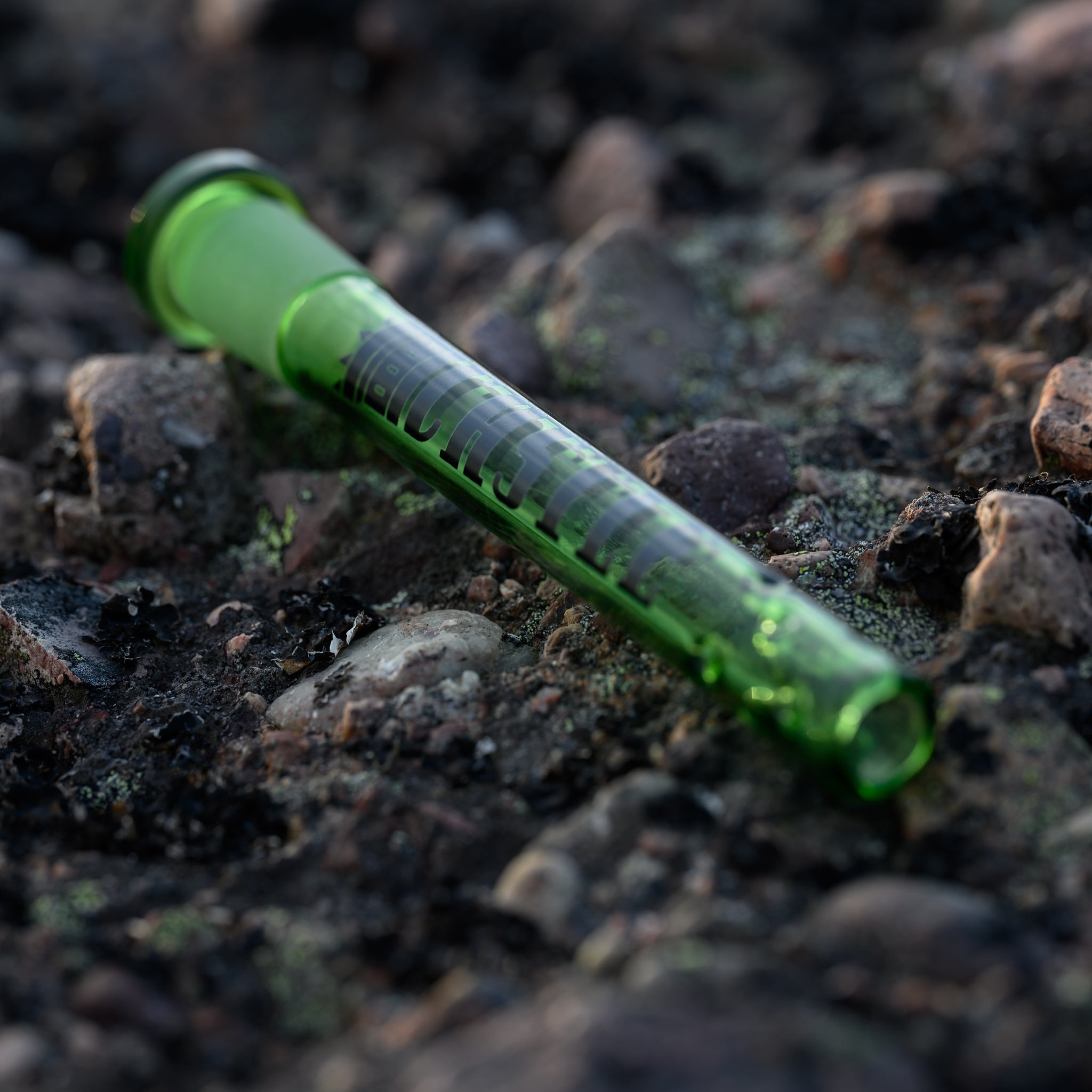 A vibrant green glass downstem with the Castle Glassworks logo is lying horizontally on a rocky, moss-covered surface. The downstem's green glass contrasts with the rough texture and earthy tones of the rocks, highlighting its sleek and polished design. The scene is naturally lit, emphasizing the downstem's smooth finish and intricate details.