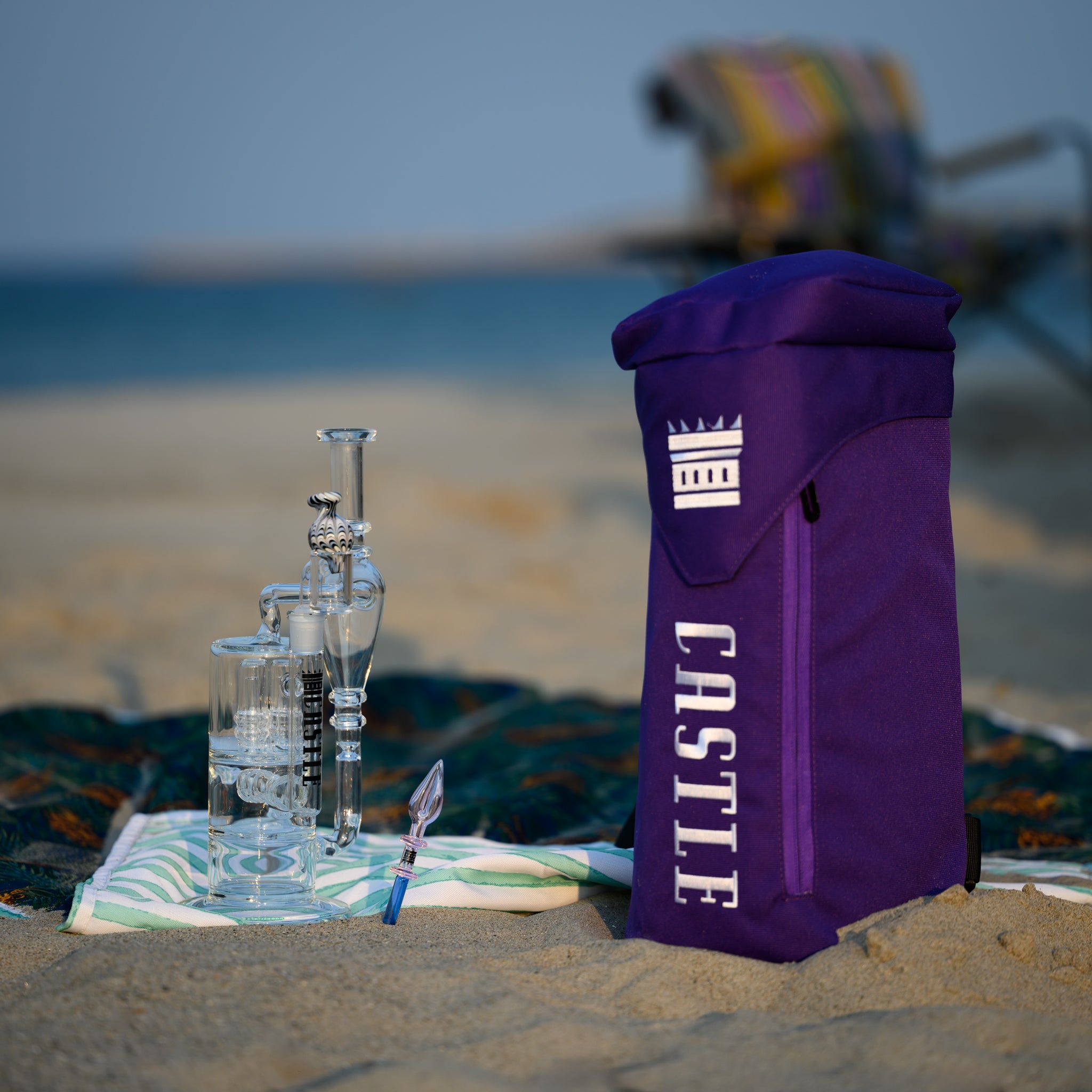 A purple bong bag with the "Castle" logo stands upright on the sand at the beach. Next to it, a clear glass rig with intricate details is placed on a colorful blanket. In the background, there is a beach chair with a colorful striped towel draped over it, suggesting a relaxed beach setting.