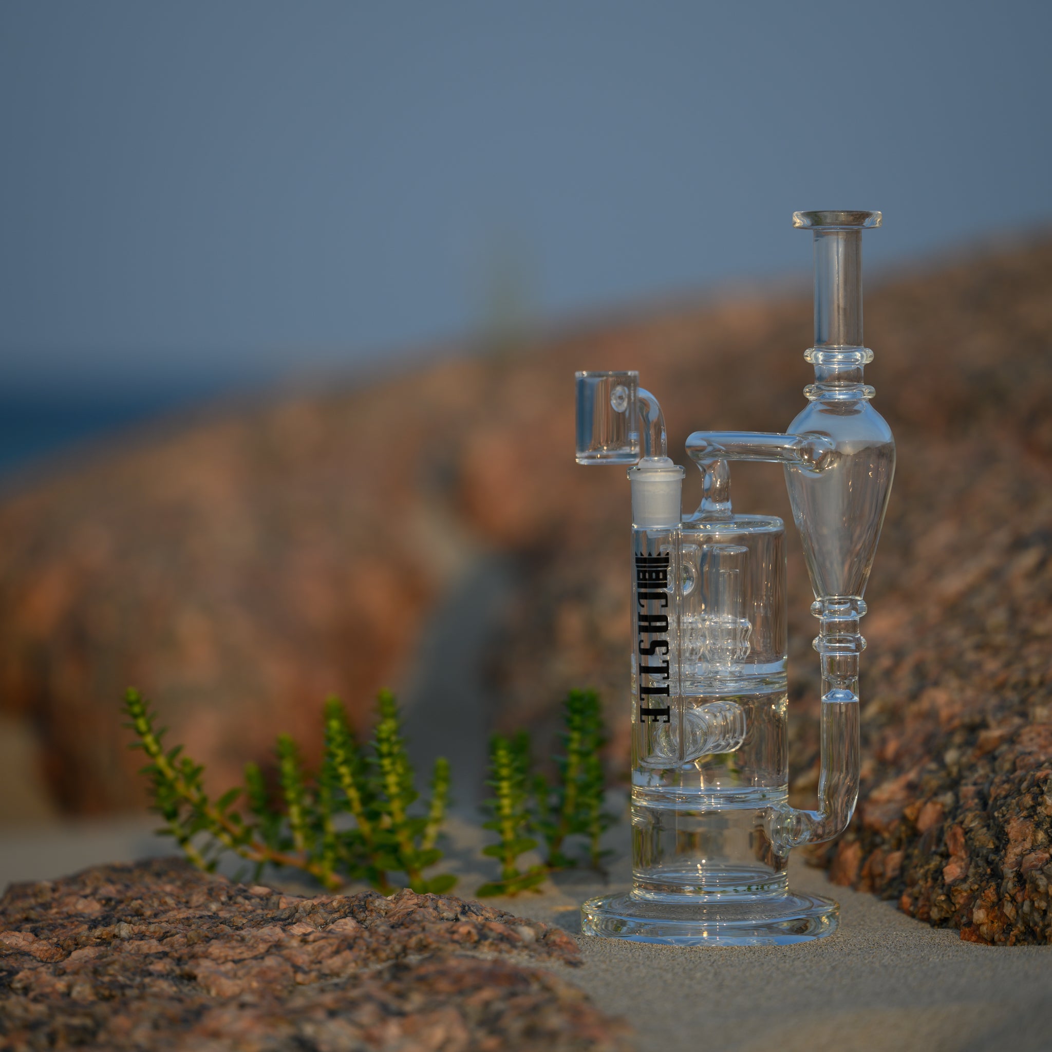 A clear glass dab rig with intricate design elements, featuring the "Castle" logo vertically. The dab rig is placed on sandy ground among rocks, with a few small green plants nearby. The natural setting includes a blurred background of rocky terrain and a hint of blue sky, creating a serene and earthy atmosphere. The sunlight enhances the transparency and craftsmanship of the glass, highlighting the detailed construction of the rig.