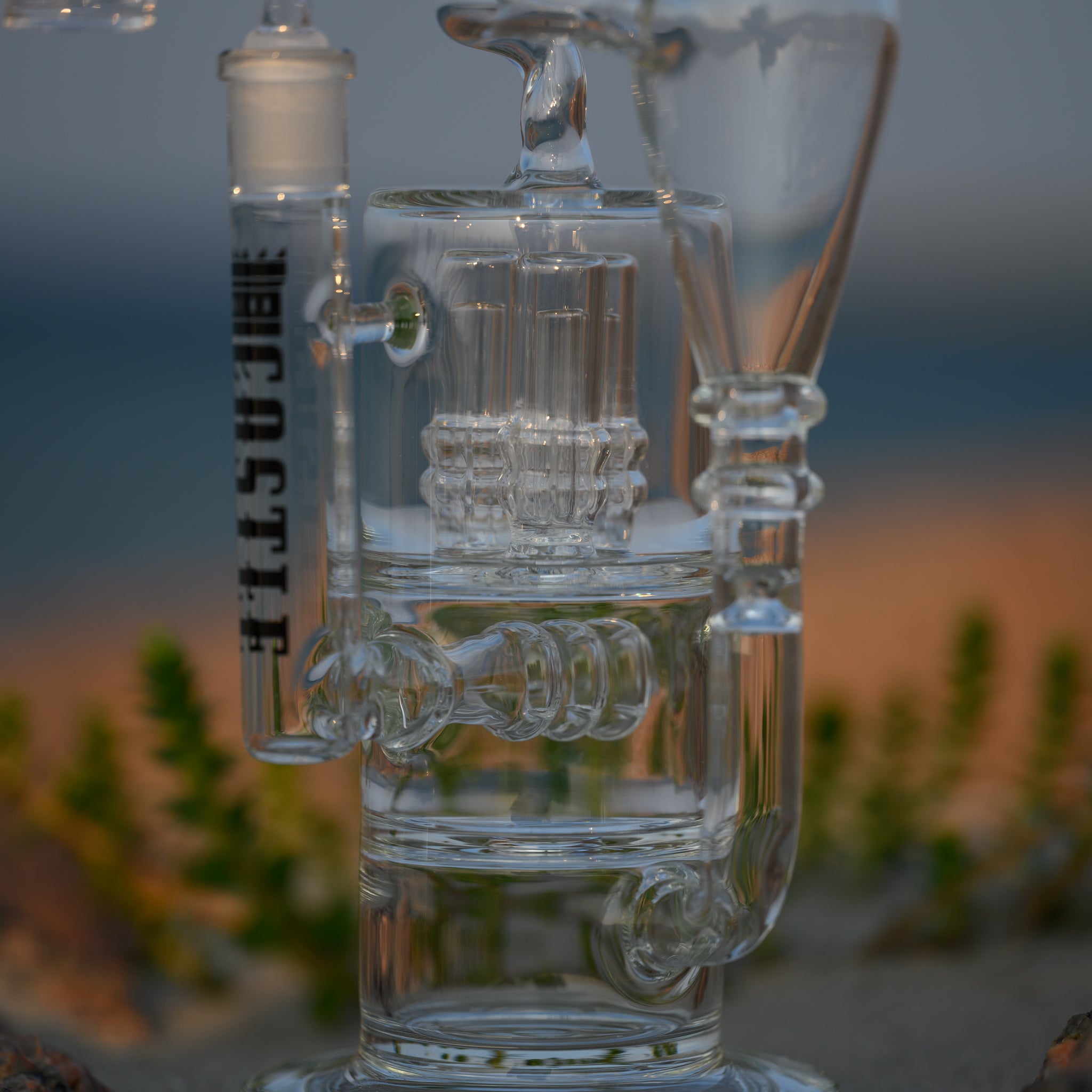 A close-up view of a clear glass dab rig with intricate design elements, featuring the "Castle" logo vertically. The detailed craftsmanship of the rig, including the complex percolators and connectors, is highlighted. The dab rig is placed on sandy ground with small green plants visible in the background. The natural setting, with the blurred rocky terrain and blue sky, creates a serene and earthy atmosphere, enhancing the beauty and transparency of the glass.