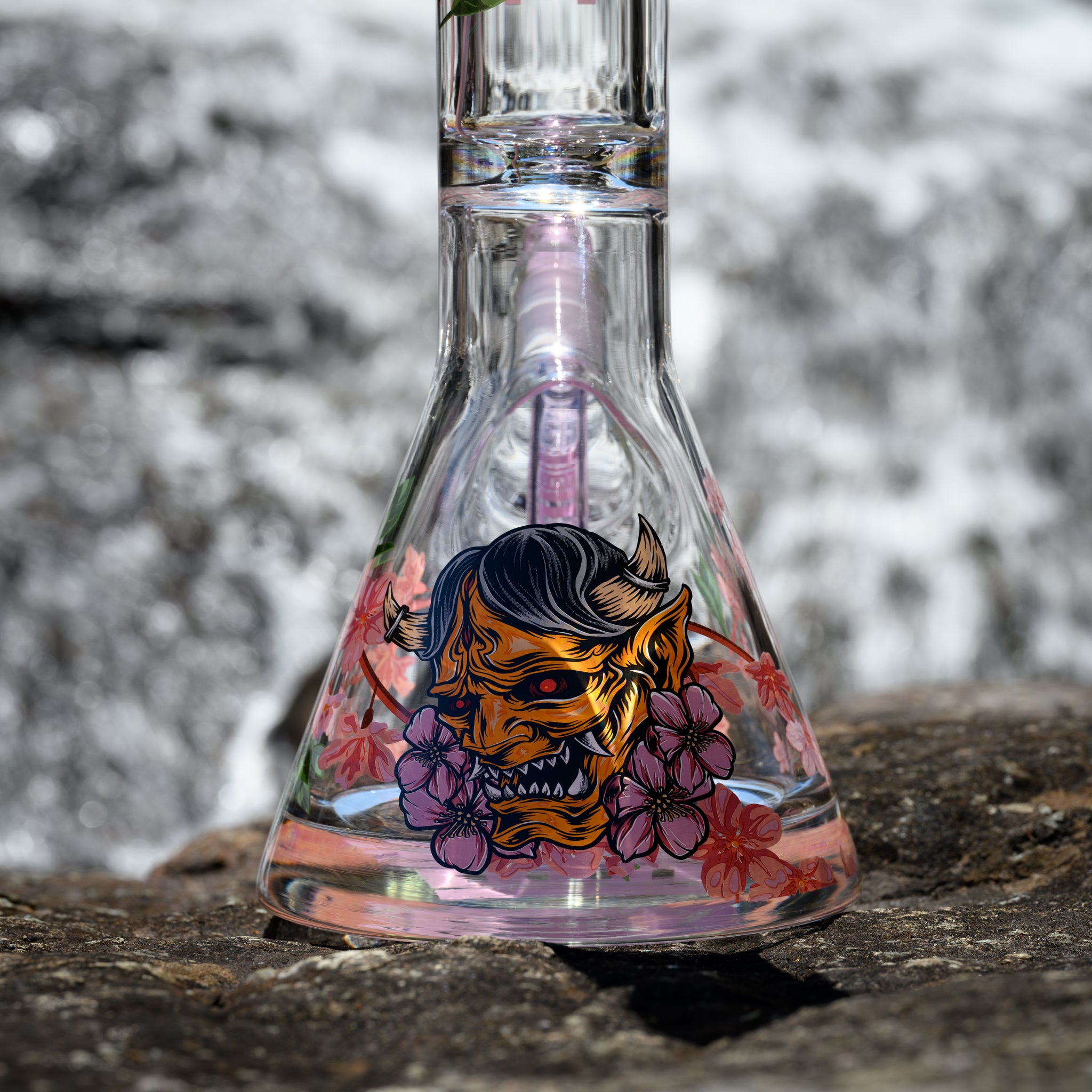 A close-up view of a clear glass beaker bong featuring a vibrant and detailed design of a traditional Japanese demon mask. The mask is surrounded by cherry blossom flowers, adding a delicate touch to the fierce image. The background showcases a blurred, flowing river, highlighting the bong's intricate artwork against the natural setting.