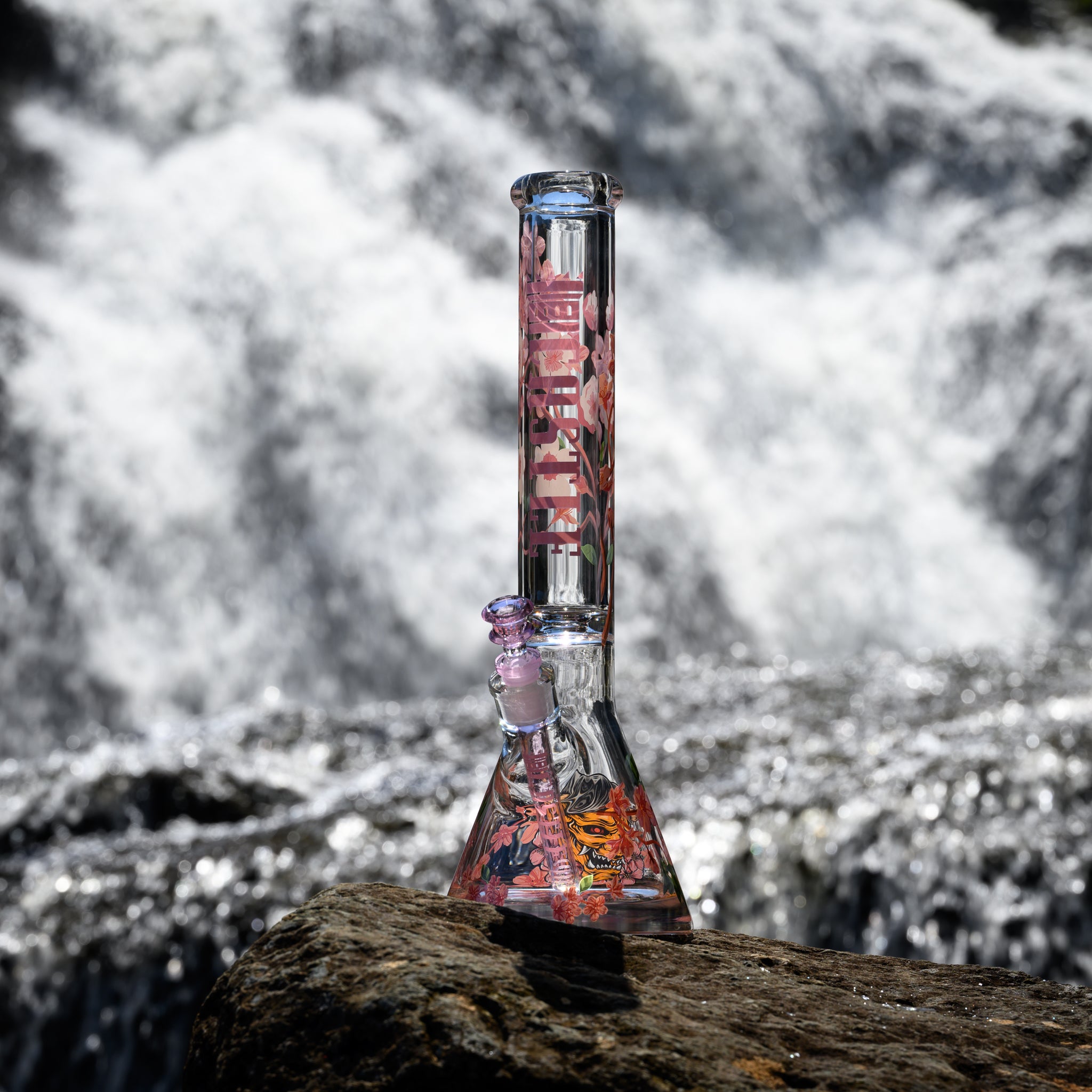 A clear glass beaker bong with cherry blossom designs stands on a rock in front of a flowing river. The intricate pink and white floral patterns on the bong contrast beautifully with the natural, dynamic backdrop of the cascading water.