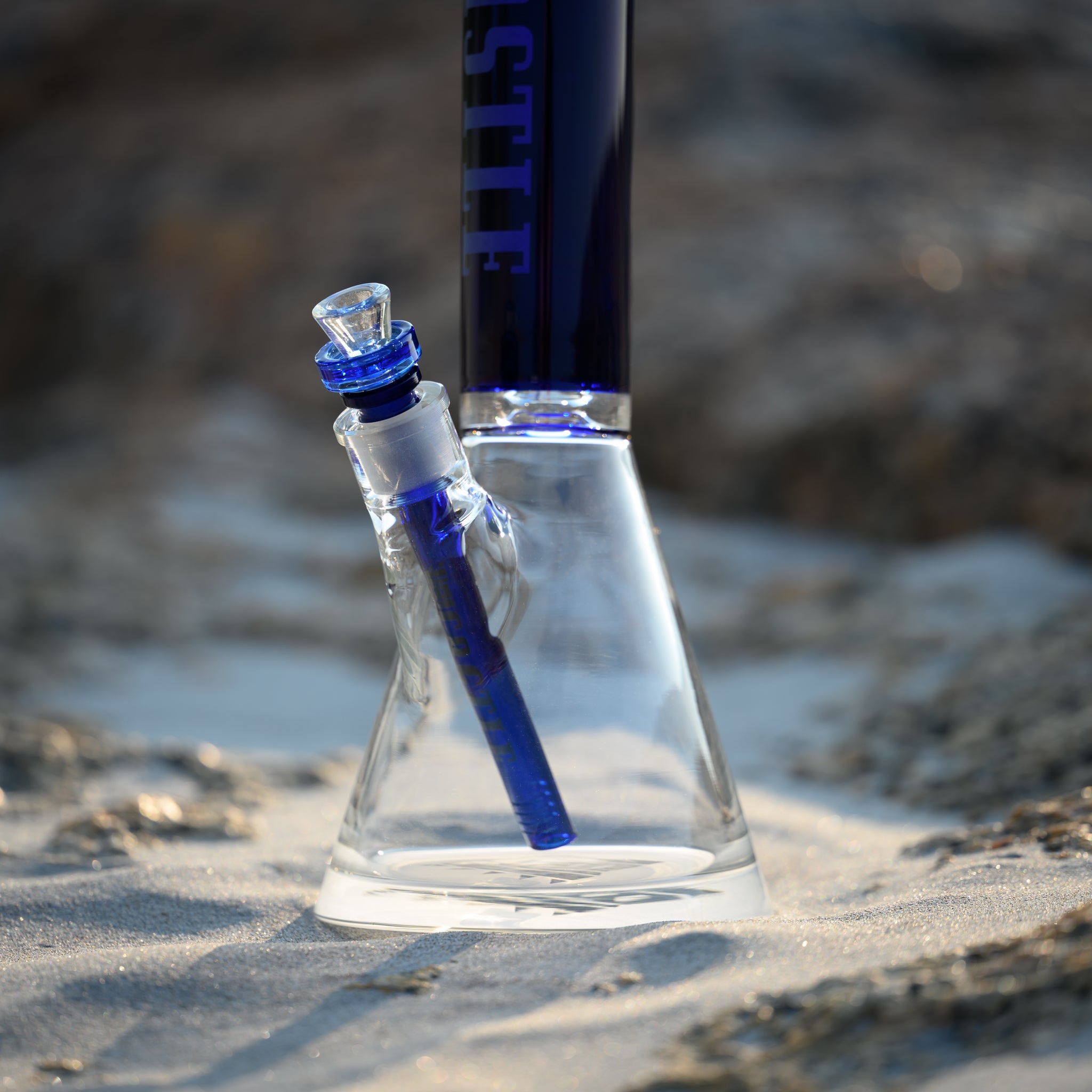 A clear glass beaker bong with deep blue accents, featuring the "Castle" logo vertically etched on the neck. The bong is positioned on sandy ground, with rocks in the background creating a natural, beach-like setting. The blue downstem and bowl complement the logo and accents, highlighting the detailed craftsmanship of the glass. The soft sunlight casts gentle shadows, enhancing the clarity and elegance of the bong while blending it harmoniously with the serene, earthy environment.