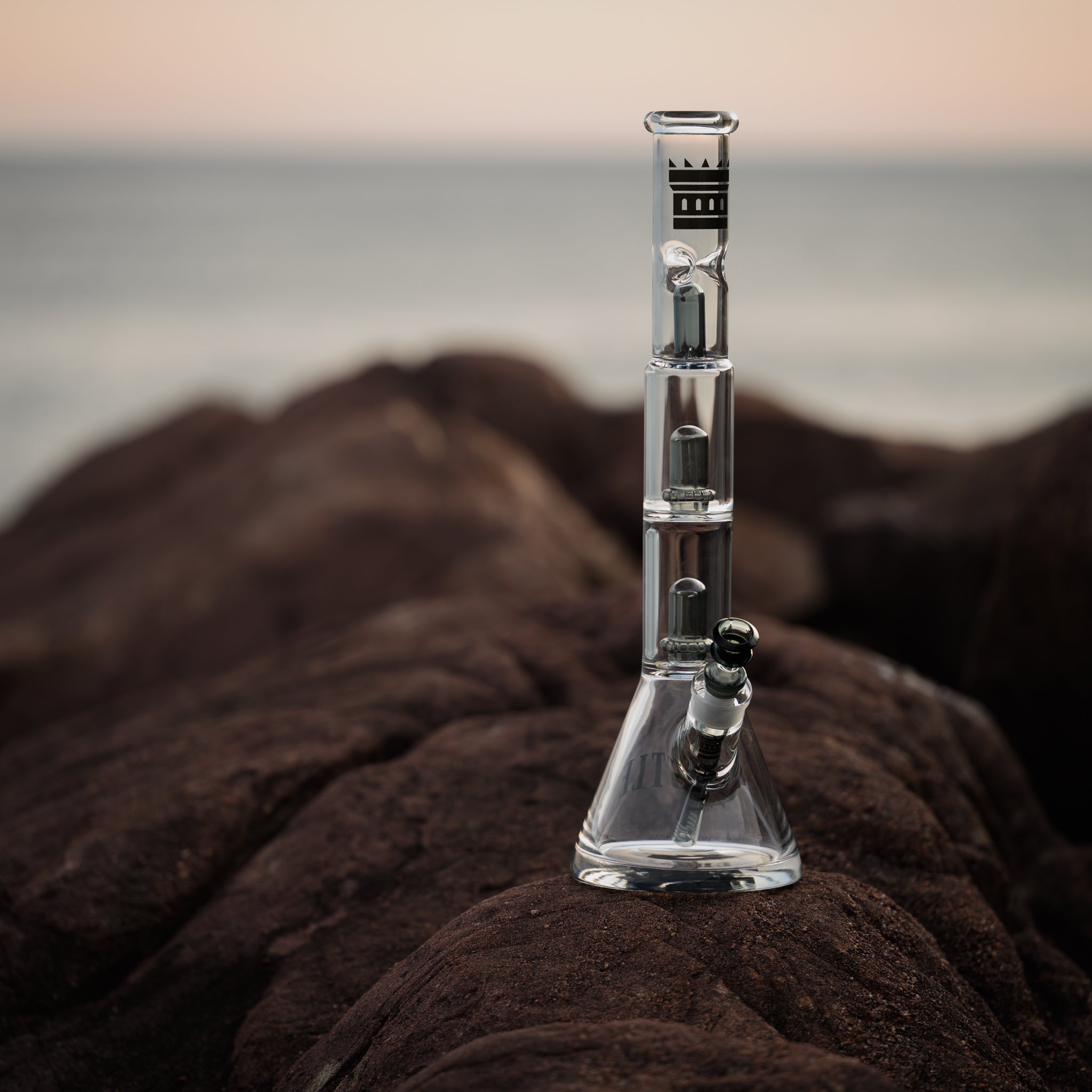 A glass bong with a double showerhead percolator stands on a rugged rock surface, with the ocean and a pastel sky visible in the background. The bong features intricate glasswork and the Castle logo, offering a striking contrast against the natural, earthy tones of the rocks and the serene seascape.