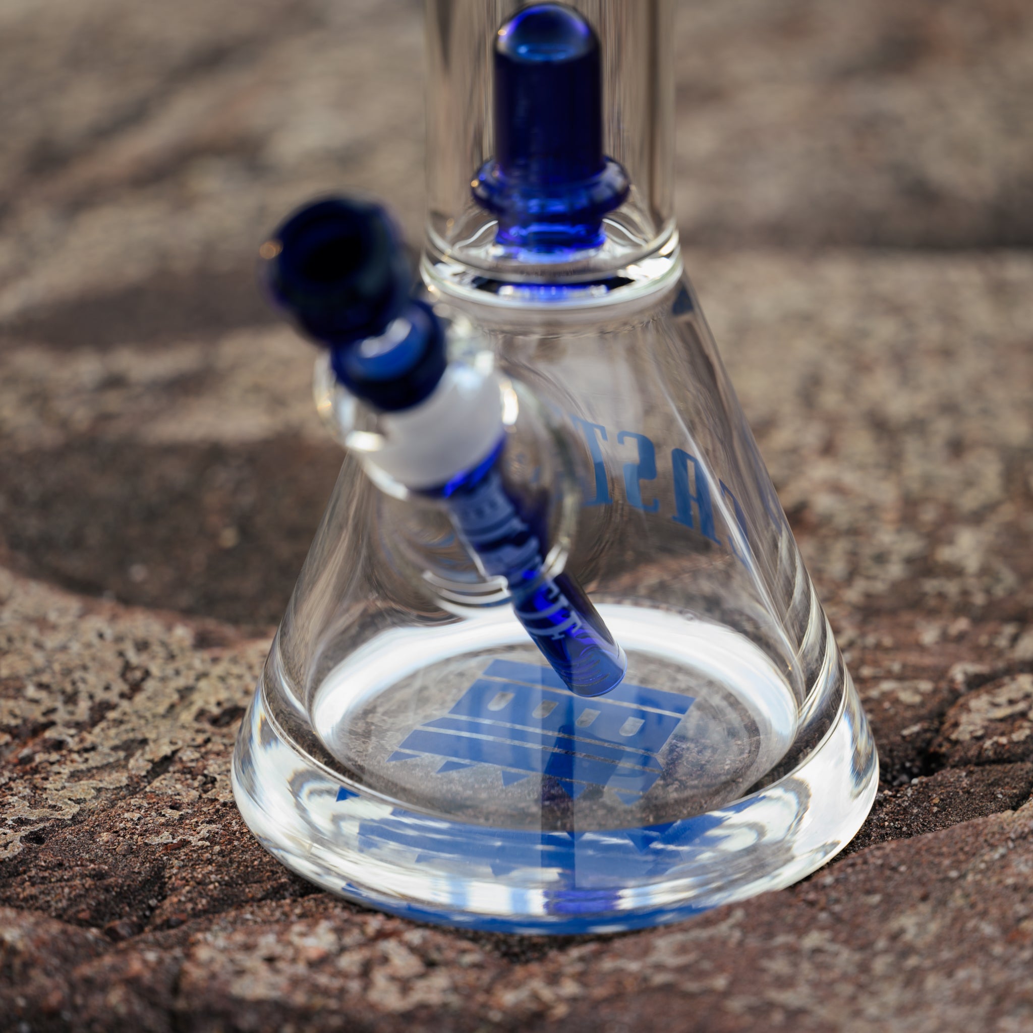 Close-up view of a glass bong with blue accents and a double showerhead percolator, standing on a textured rock surface. The base of the bong features the Castle logo in blue, with intricate glasswork visible through the transparent body. The blue downstem and bowl add a vibrant touch to the overall design.