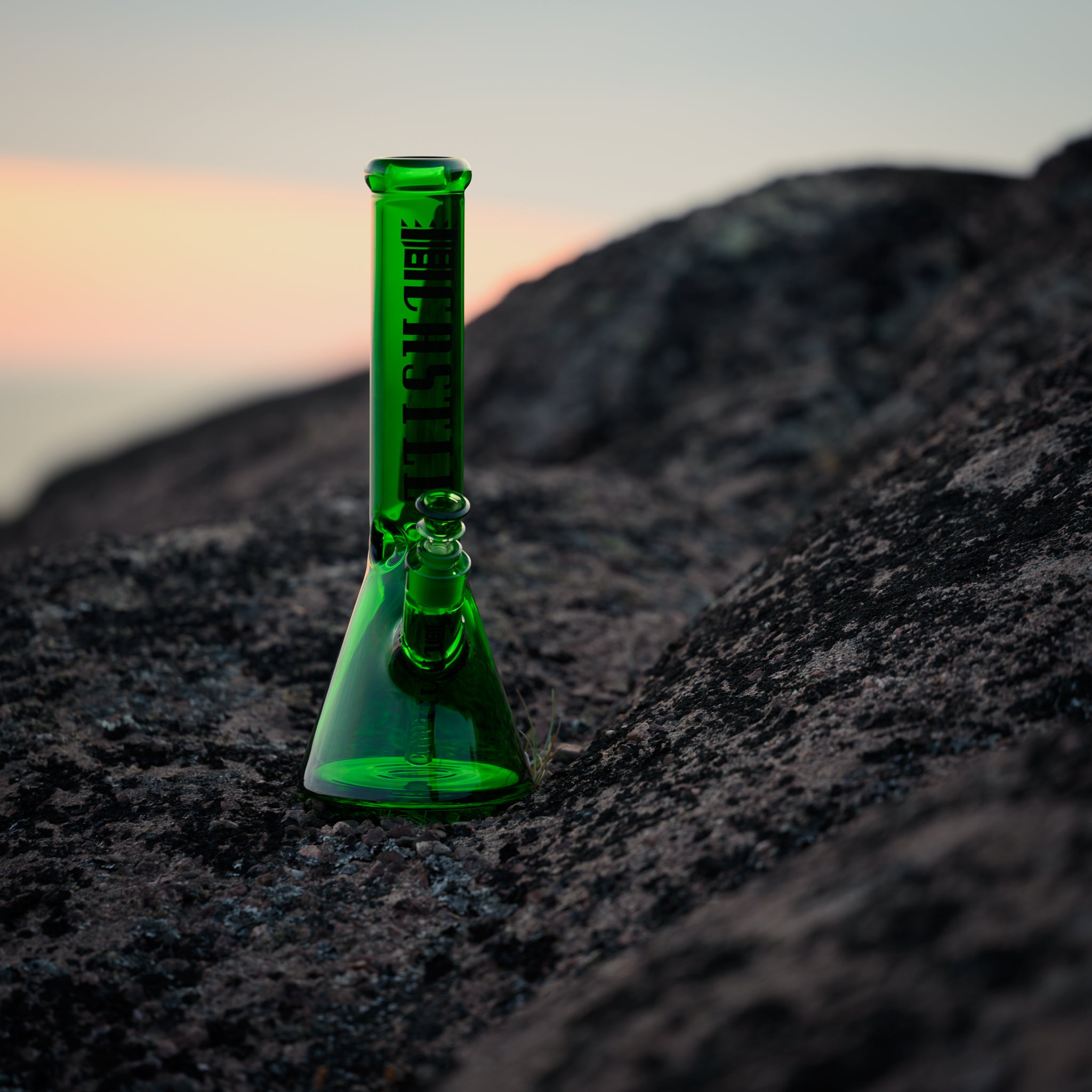 A vibrant green electroplated beaker bong with Castle Glassworks branding is placed on a rocky surface during sunset. The green glass contrasts sharply with the dark, textured rock and the soft pastel colors of the sky in the background, creating a striking visual effect. The lighting accentuates the smooth finish and transparency of the bong, highlighting its intricate design and the brand's logo.