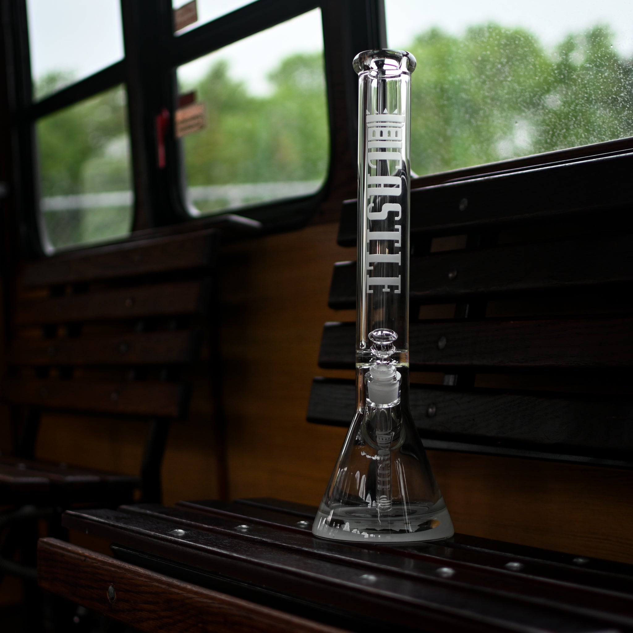 A clear glass beaker bong with the "Castle" logo etched vertically on the neck, featuring sleek and modern design elements. The bong is positioned on a wooden bench inside a vintage bus. The interior of the bus includes wooden slat seats and windows with a view of greenery outside, slightly blurred and dotted with raindrops. The scene combines the classic charm of the bus's interior with the contemporary aesthetic of the bong.