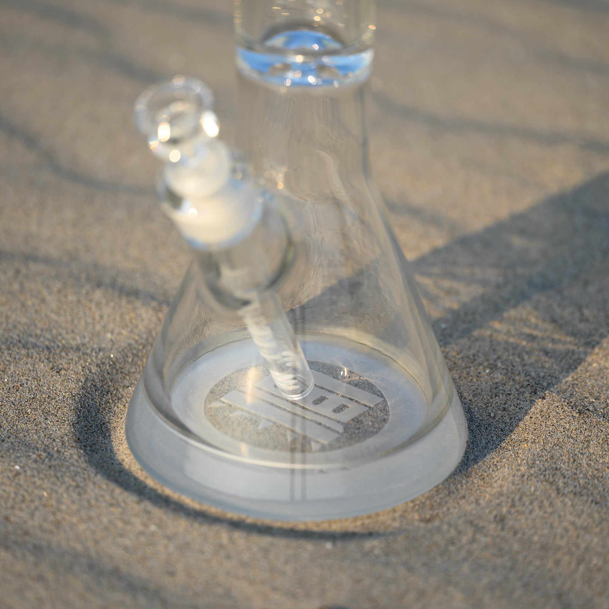 A close-up view of a clear glass beaker bong with an etched "Castle" logo on the base, positioned on sandy ground. The downstem and bowl are visible, with soft sunlight casting gentle shadows across the sand. The clarity and craftsmanship of the glass are highlighted, creating an elegant and serene atmosphere. The etched logo adds a touch of sophistication to the minimalist and natural setting.