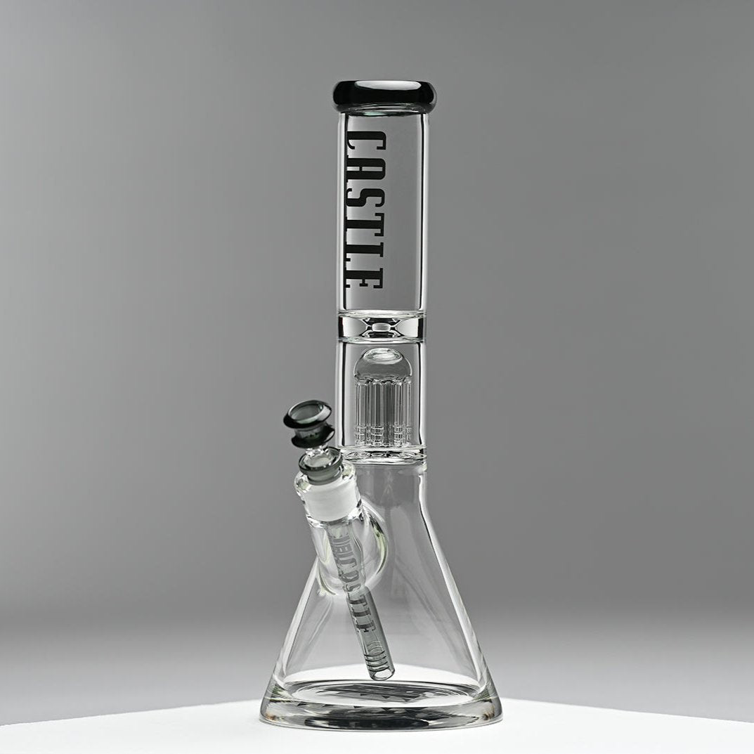 A glass beaker bong with the "Castle" logo prominently displayed on the neck, featuring an 8-arm percolator inside. The bong is set against a minimalist grey background, emphasizing its clean lines and modern design. The photo captures the bong from the left side, highlighting its structure and details.