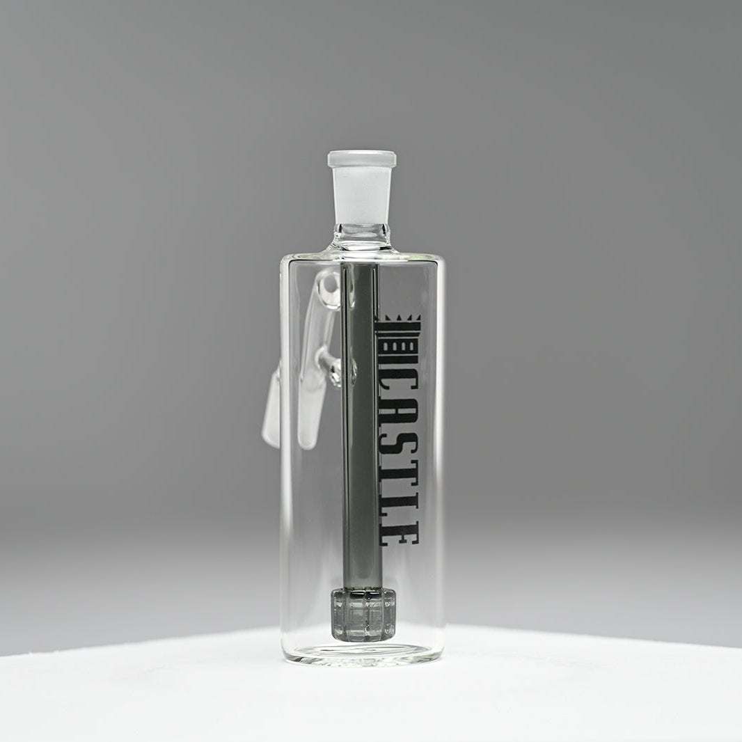 A glass ash catcher with a modern and minimalist design, featuring the brand name Castle in bold black letters on the side. The ash catcher includes a black matrix percolator, adding both aesthetic appeal and functionality. The piece is set against a neutral grey background, which emphasizes the clarity and sleekness of the glass.