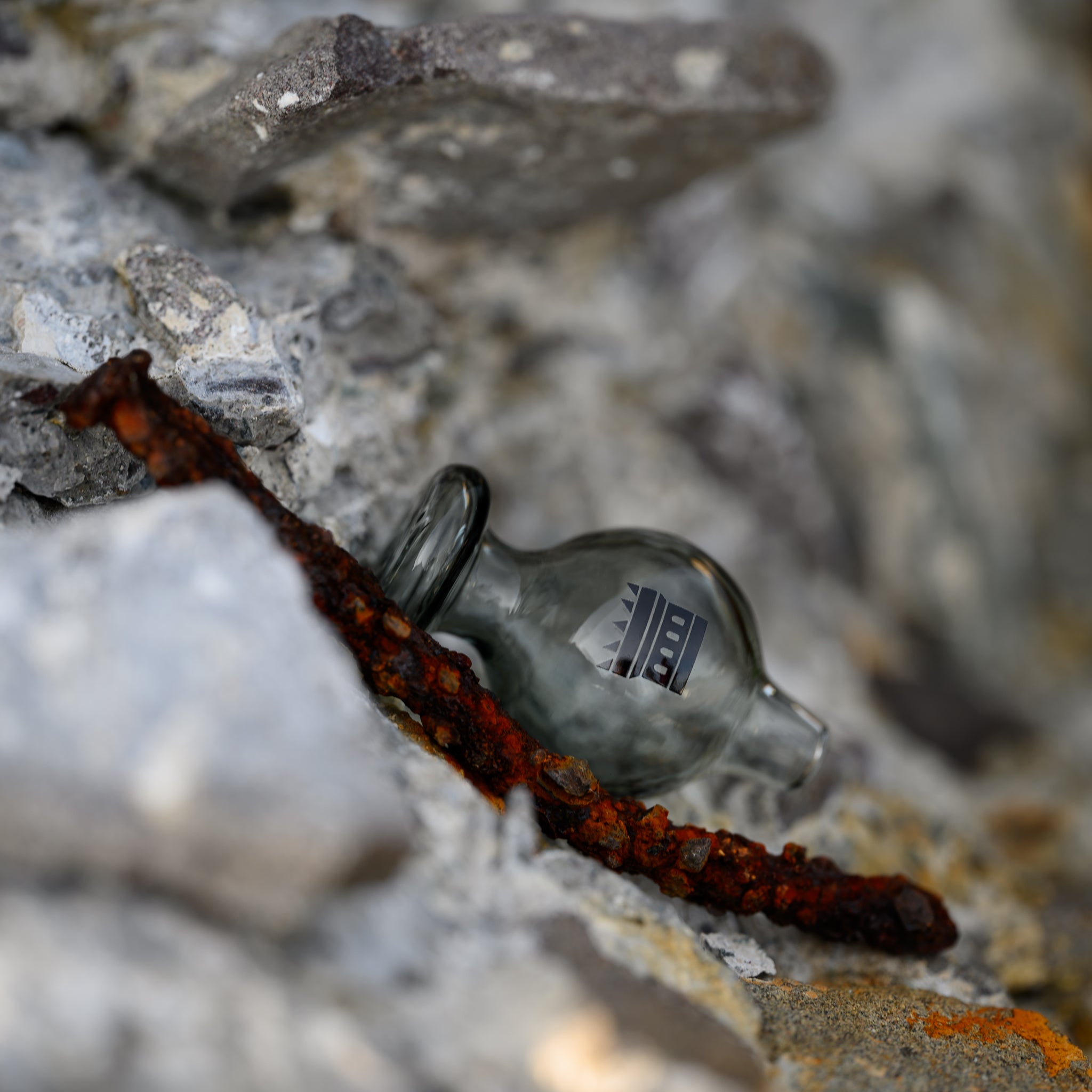 A grey glass carb cap with the Castle Glassworks logo rests against a backdrop of rugged stone and a piece of rusty metal. The contrast between the smooth, modern design of the carb cap and the rough, weathered textures of the surrounding elements creates a striking visual effect.