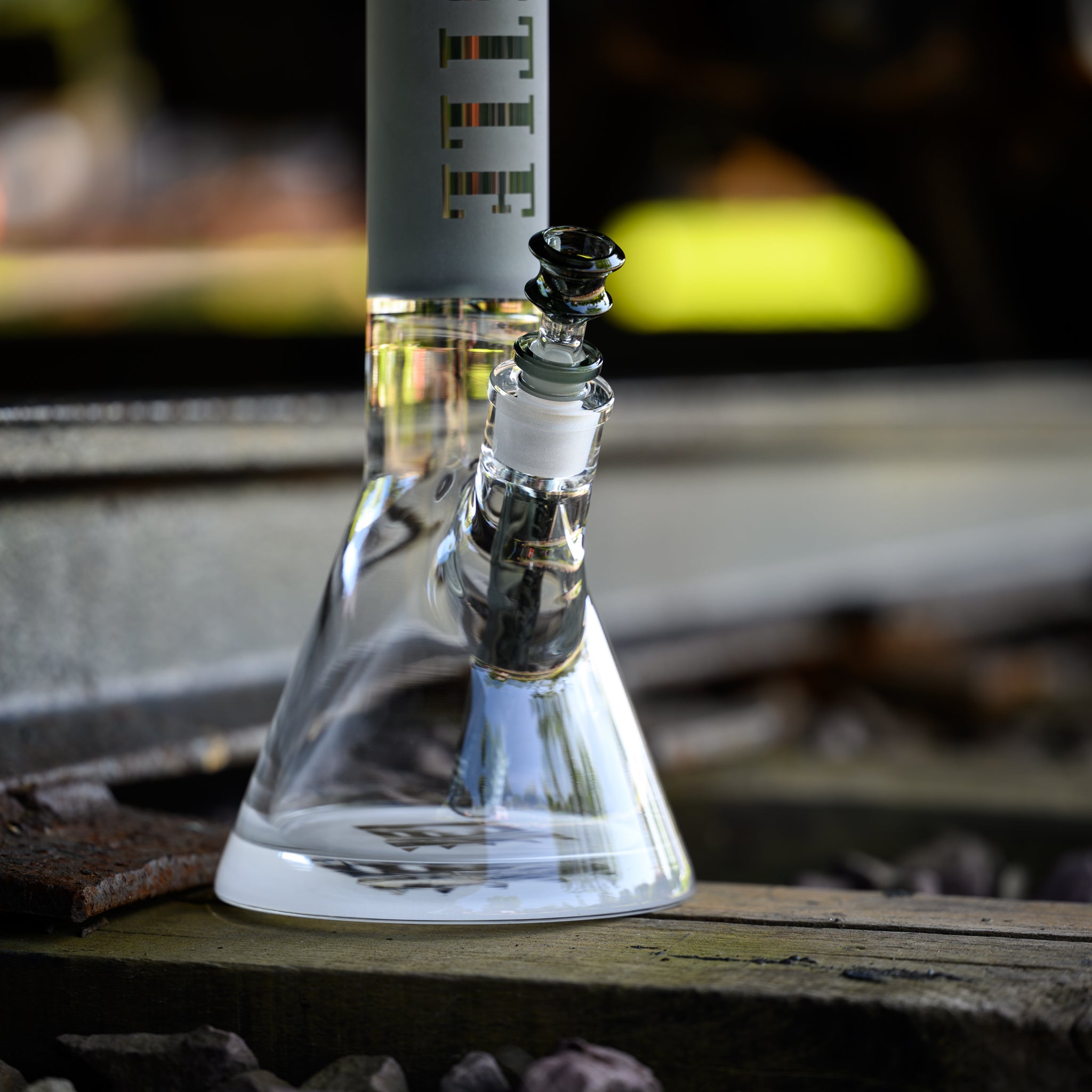 A close-up view of a clear glass beaker bong with a frosted grey neck, featuring the "Castle" logo etched vertically. The bong is positioned on a wooden plank by a railway track, with gravel and metal rails visible in the background. The downstem and bowl, made of clear and black glass, are prominently displayed. The scene blends industrial elements with the sleek design of the bong, creating a mix of textures and themes.