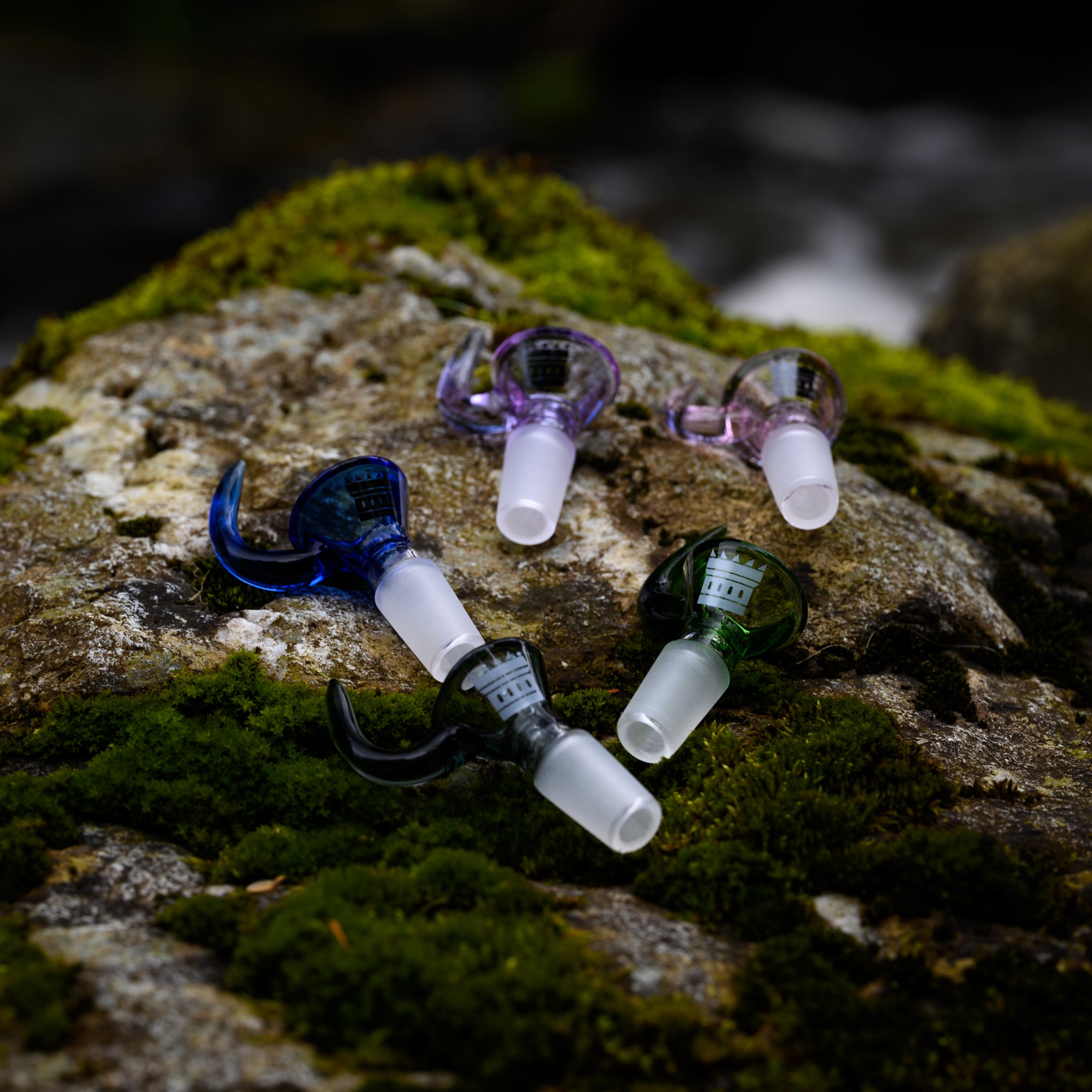 A collection of colorful glass bong bowls with horn-like handles, displayed on a moss-covered rock. Each bowl features a frosted joint and a distinctive horn design, adding a touch of elegance and uniqueness to their appearance. The natural setting of the mossy rock and the blurred background of flowing water create a serene and earthy atmosphere, highlighting the contrast between the crafted glass and the organic environment.