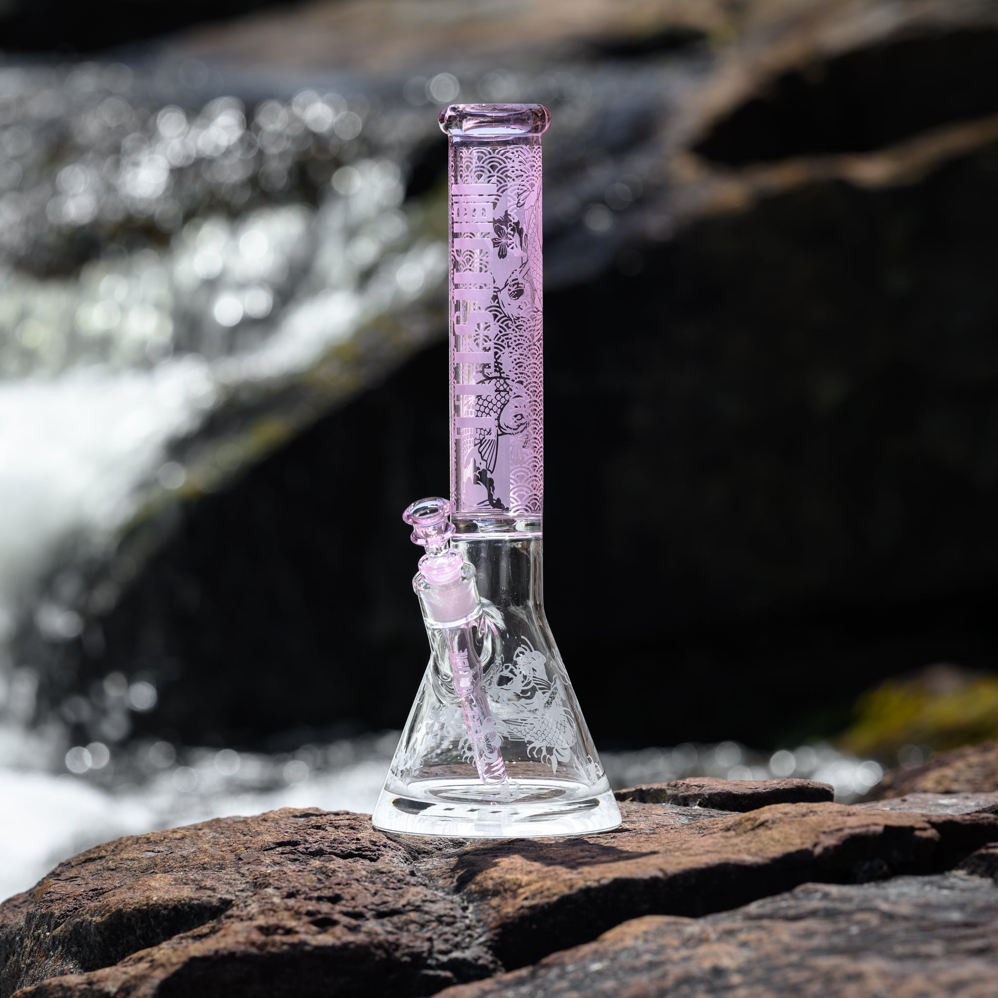 A glass beaker bong with a frosted pink neck and the "Castle" Logo vertically on the neck, adorned with intricate designs featuring koi fish and Japanese-style wave patterns. The downstem and bowl also feature matching pink accents. The bong is placed on a rocky surface beside a flowing river, with the water creating a sparkling, blurred background. The detailed artwork on the bong is highlighted by the natural light, emphasizing the craftsmanship and the serene outdoor setting.