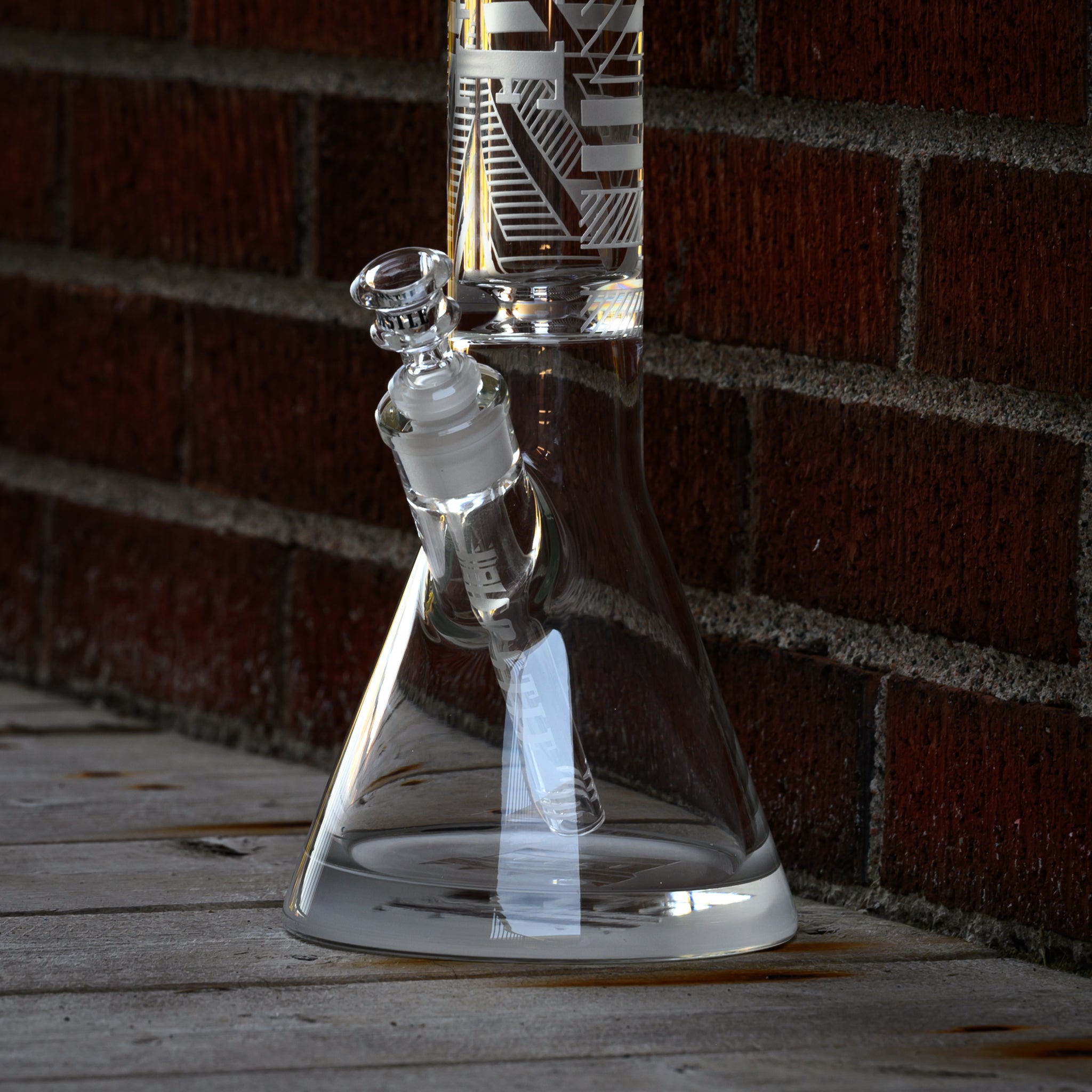  close-up view of a clear glass beaker bong with geometric designs etched onto its surface, positioned against a brick wall. The downstem and bowl are clearly visible, emphasizing the precision and clarity of the glass craftsmanship. The setting includes a wooden floor, adding to the rustic and urban feel of the scene. The combination of the modern, sleek design of the bong with the textured brick and wood backdrop creates an interesting contrast and visual appeal.