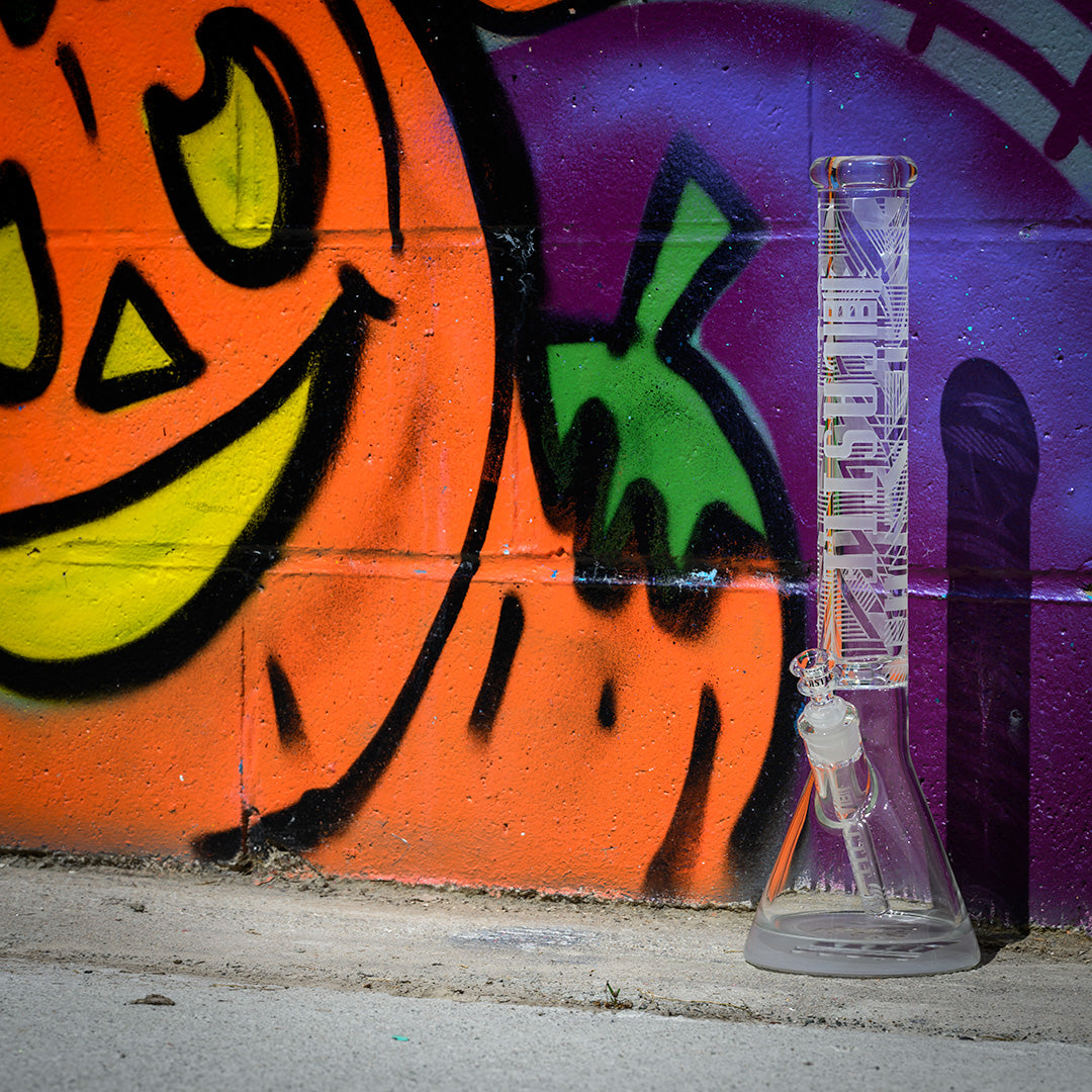 A clear glass beaker bong with geometric designs etched onto its surface, standing against a vibrant graffiti wall. The graffiti features a large, colorful pumpkin with a happy face, painted in bold orange, yellow, green, and purple hues. The sleek, modern design of the bong contrasts with the playful and artistic graffiti, creating a dynamic and visually engaging scene. The concrete ground and brick wall add an urban feel to the image.