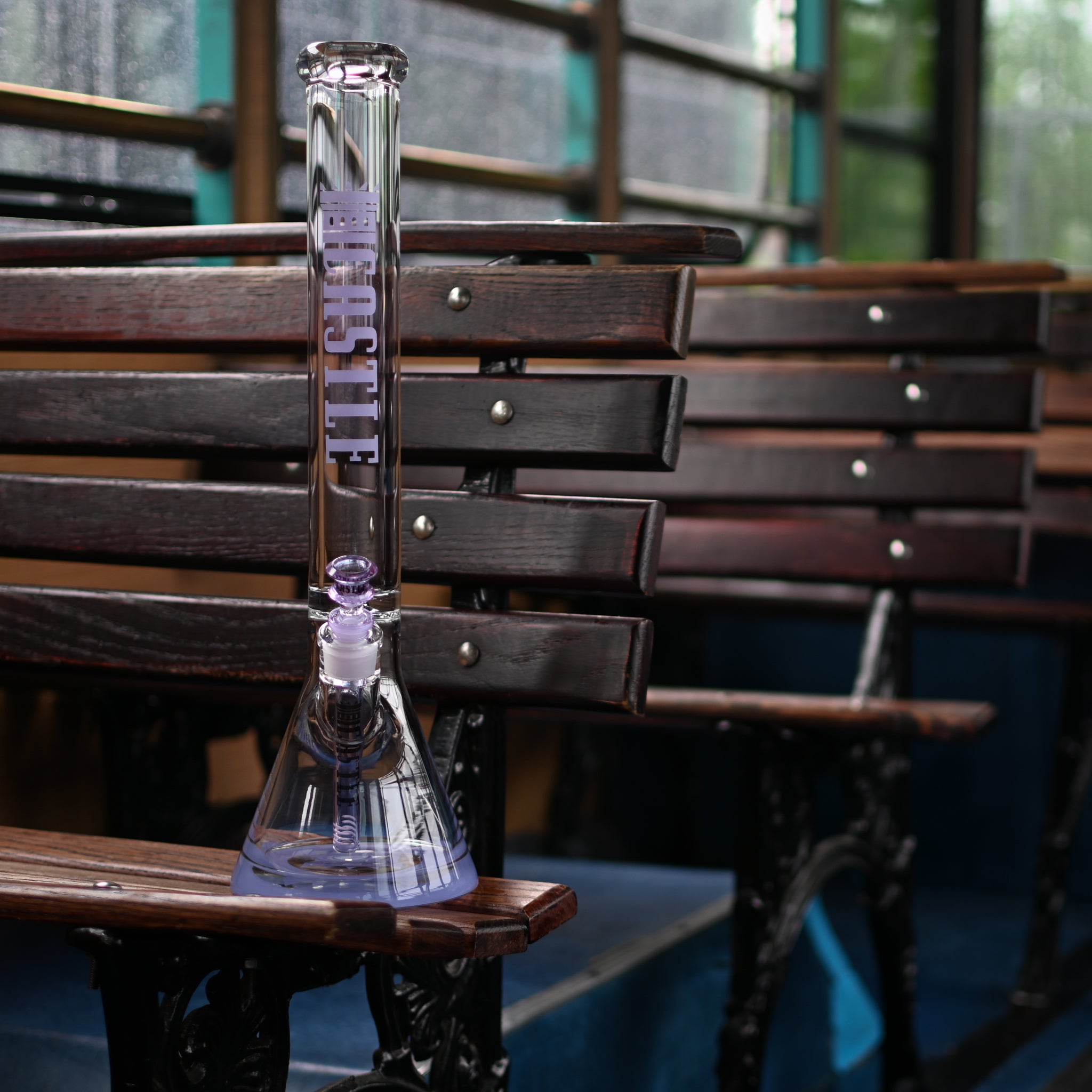 A clear glass beaker bong with purple accents and the "Castle" logo vertically displayed on the neck, positioned on a wooden bench inside a vintage bus. The wooden slats of the bench contrast with the sleek glass, and the background shows more benches and the bus's interior, featuring blue and teal tones. The lighting is soft, with natural light filtering through the bus windows, creating a cozy and nostalgic ambiance.