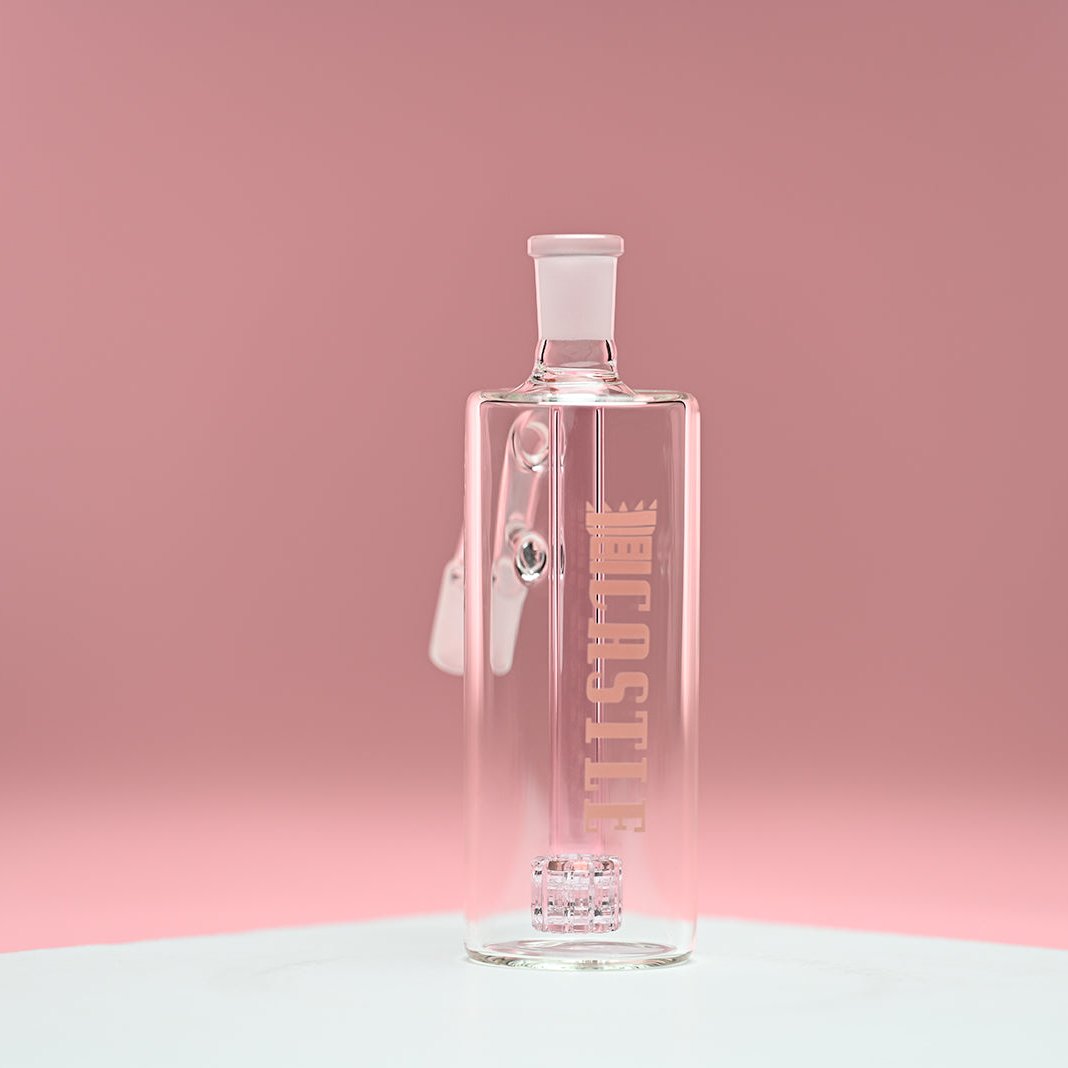 A glass ash catcher featuring the brand name Castle in soft pink letters on the side, with a matching pink matrix percolator. The ash catcher is positioned against a smooth pink background, which enhances the delicate pink color of the percolator and branding. The transparent glass design and precise craftsmanship are highlighted, giving the piece a modern and elegant appearance.