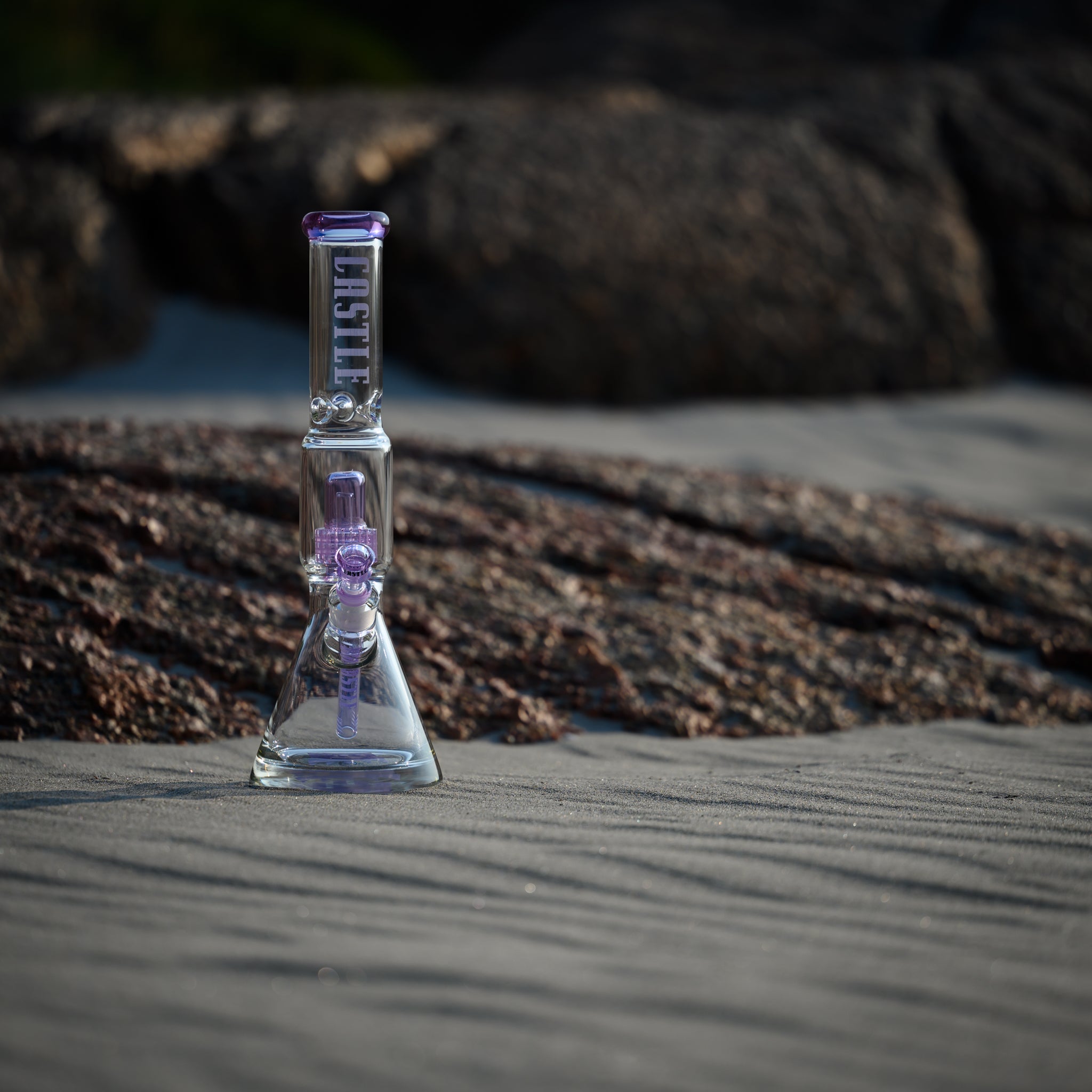 A clear glass beaker bong with purple accents, featuring the "Castle" logo, placed on sandy ground. The background includes large rocks, adding a natural and rugged contrast to the smooth and transparent glass of the bong. The sunlight casts gentle shadows, highlighting the details of the glass and the texture of the sand.