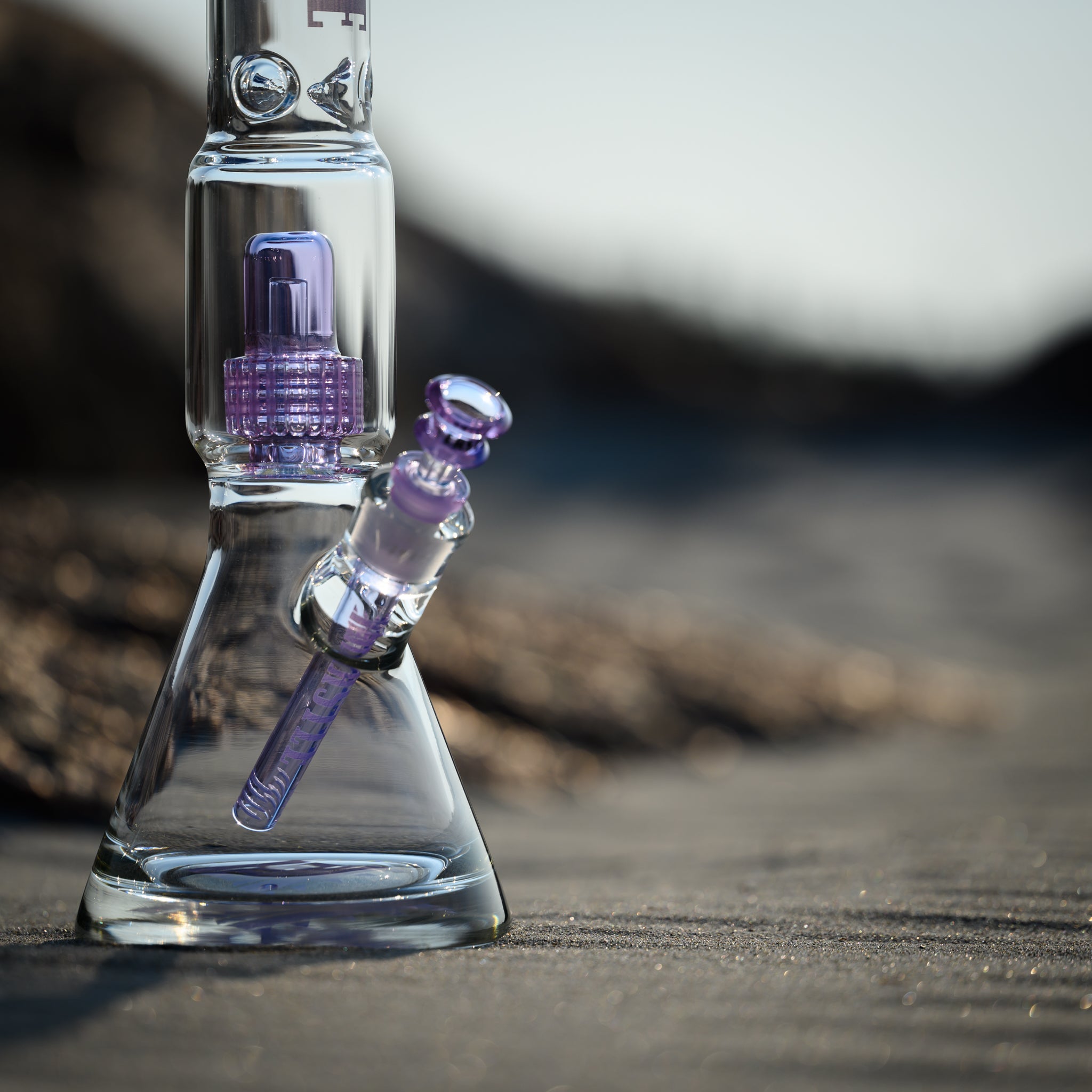 Close-up view of a glass beaker bong with purple accents and the "Castle" logo, placed on sandy ground. The image focuses on the intricate details of the bong's components, including the bowl and percolator. The background is blurred, highlighting the smooth glass against the sandy texture. The sunlight enhances the clarity and color of the purple accents.