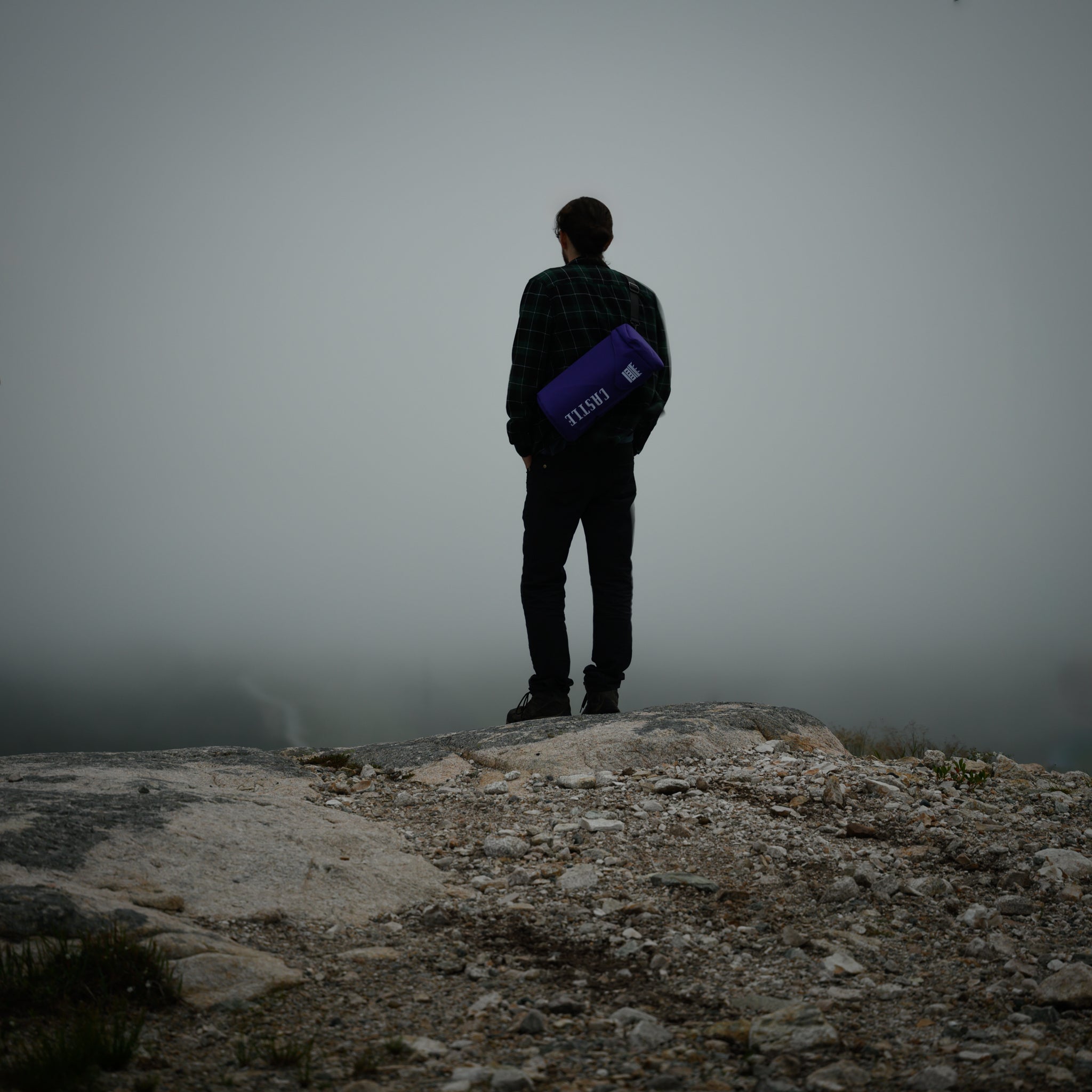 A person stands on rocky terrain, facing away and looking into a foggy distance. They are wearing a green and black checkered shirt and black pants, holding a purple bong bag with the "Castle" logo. The overall scene conveys a sense of solitude and contemplation in a misty, rugged landscape.