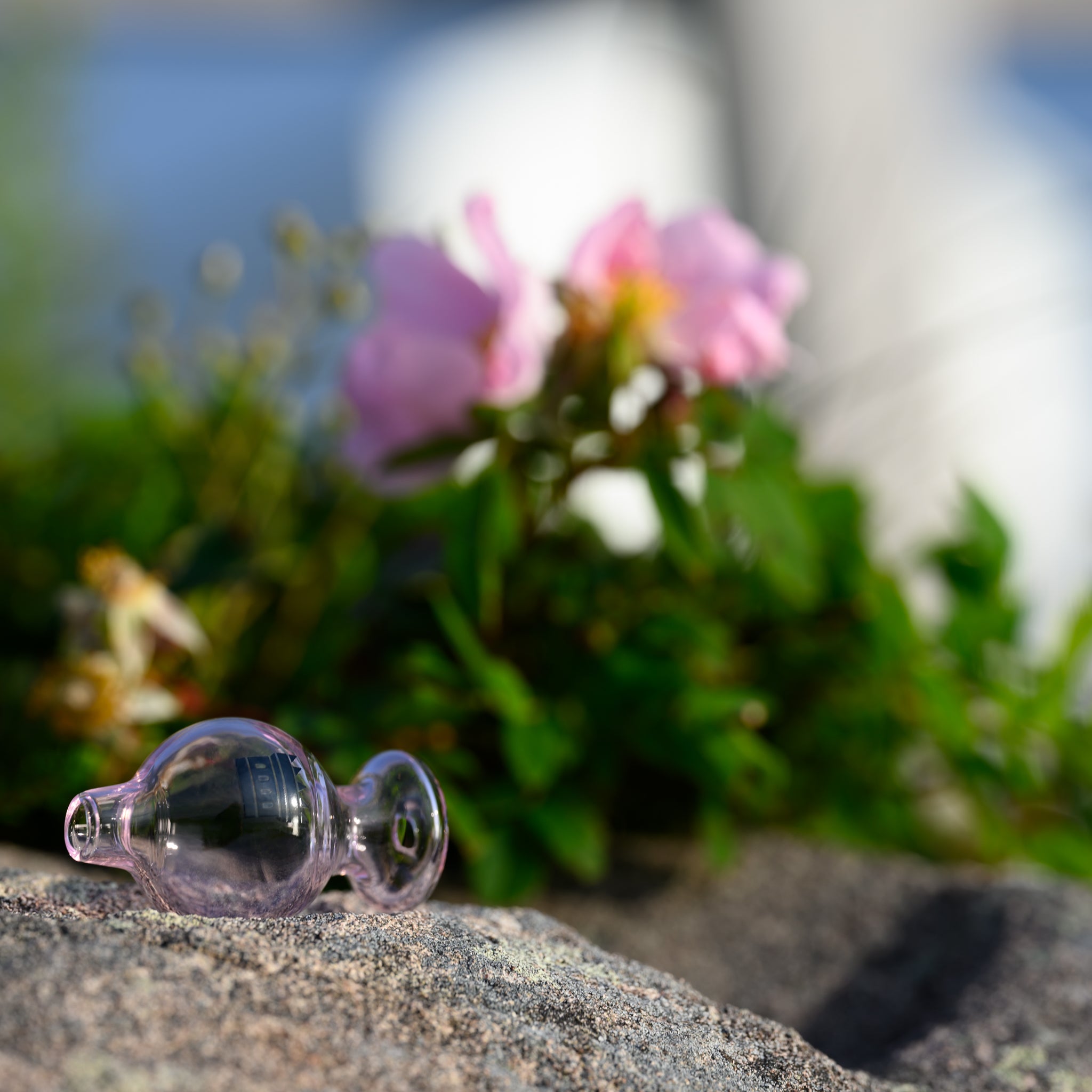 A pink glass carb cap with the Castle Glassworks logo sits on a stone surface. In the background, pink flowers and green foliage are slightly out of focus, creating a natural and serene setting that highlights the carb cap's delicate color and craftsmanship.