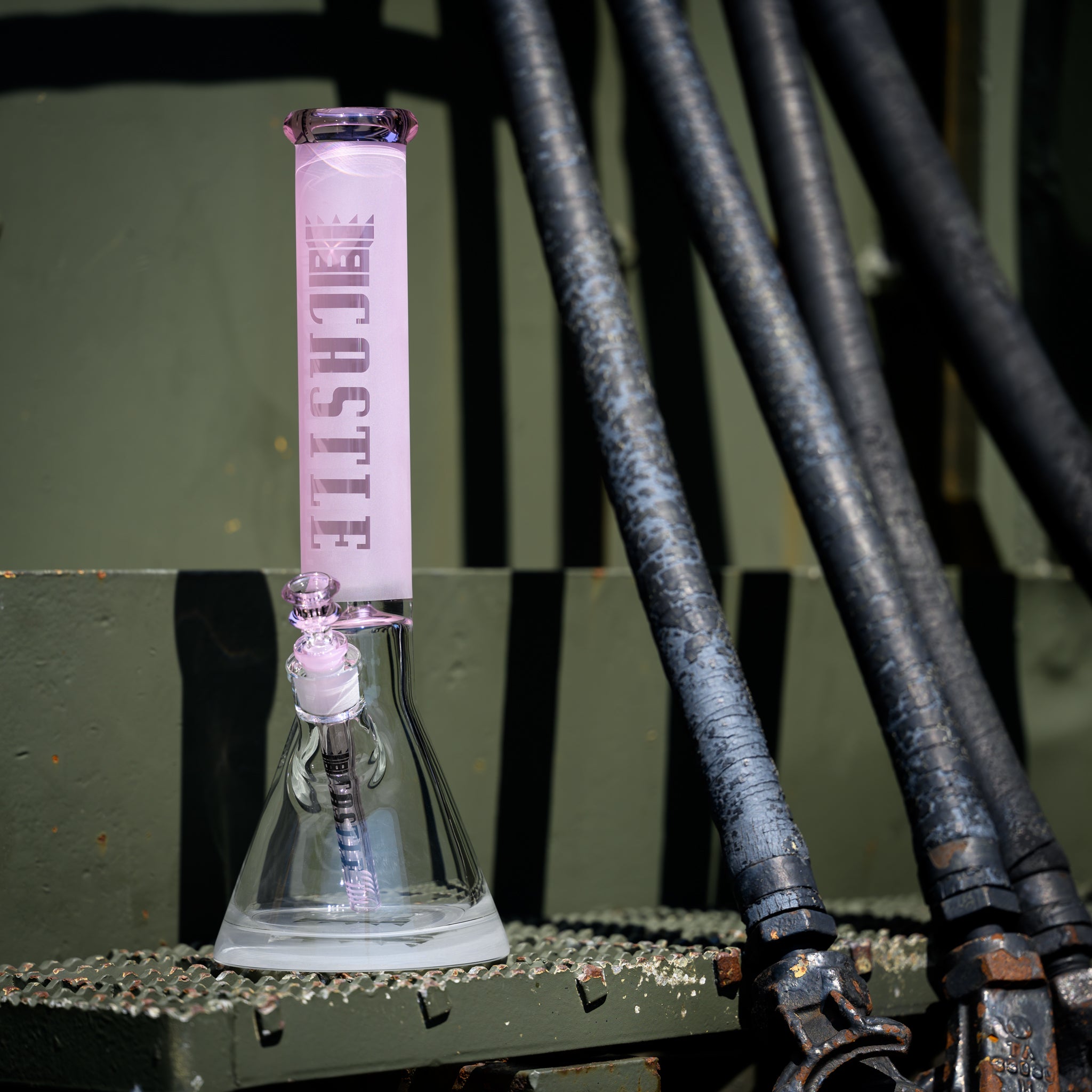 A glass beaker bong with a frosted pink neck, featuring the "Castle" logo etched vertically. The bong is placed on a textured metal surface, with several black industrial pipes running vertically in the background. The setting appears to be an industrial or mechanical environment, creating a stark contrast between the soft, pastel pink of the bong and the rugged, utilitarian backdrop. The sunlight casts sharp shadows, adding depth and emphasis to the scene.