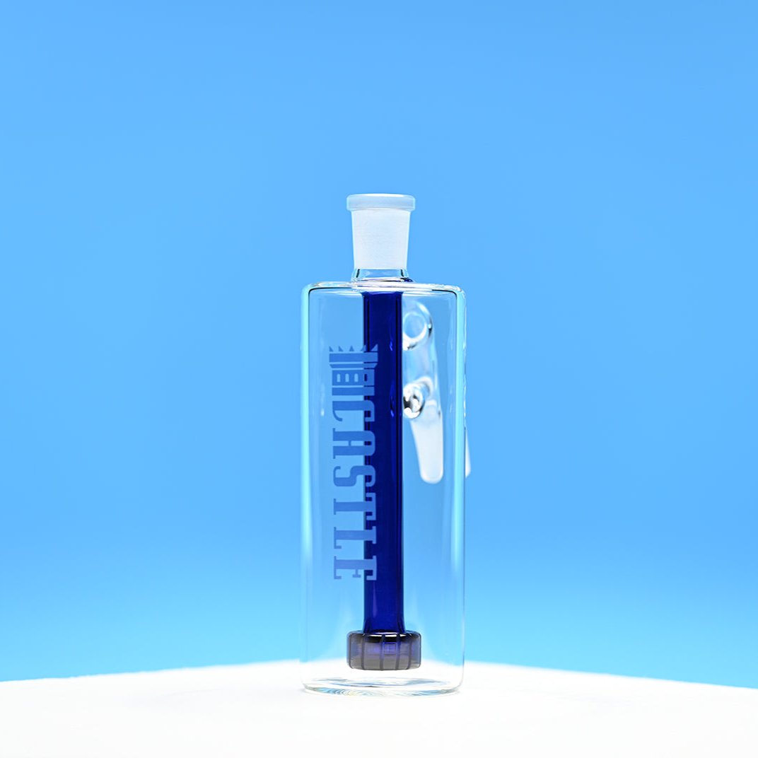 The image shows a clear glass ash catcher with a dark blue showerhead percolator inside. The ash catcher has the brand name "CASTLE" vertically on the front. The background is a solid, vibrant blue, which contrasts sharply with the dark blue percolator inside the ash catcher. The ash catcher is placed on a white surface, and the photo is taken from a front-facing angle, emphasizing the clarity of the glass and the deep color of the percolator.