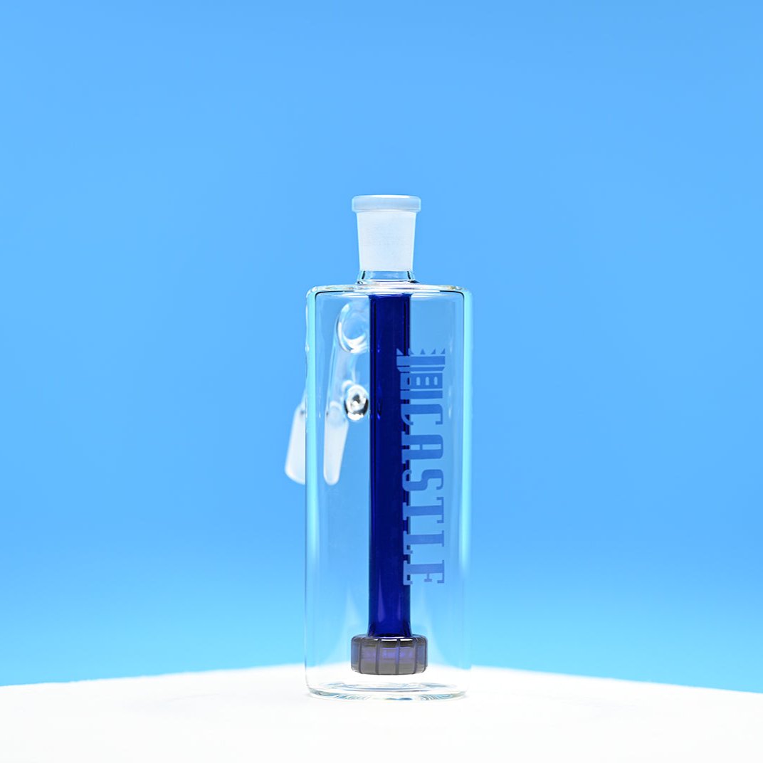 The image features a clear glass ash catcher with a dark blue showerhead percolator inside. The brand name "CASTLE" is etched vertically on the front of the ash catcher. The background is a solid, bright blue, which contrasts well with the dark blue percolator inside. The ash catcher is positioned on a white surface and photographed from a front-facing angle, showcasing its clean lines and the deep color of the percolator.