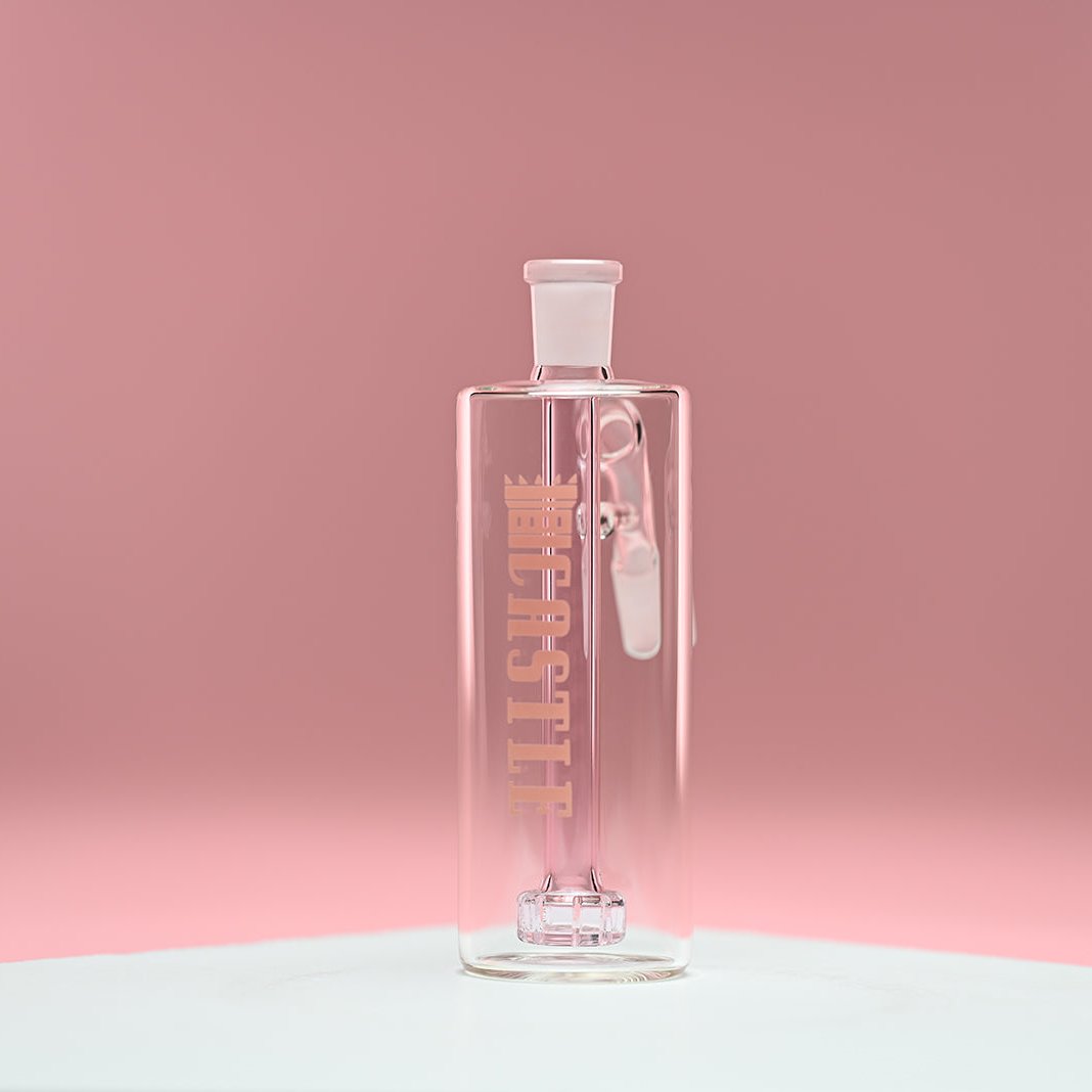 The image shows a clear glass ash catcher with a light pink showerhead percolator inside. It has the brand name "CASTLE" vertically on the front of the ash catcher. The background is a soft pink, which complements the delicate pink hue of the percolator inside. The ash catcher is positioned on a white surface and photographed from a front-facing angle, highlighting its transparent design and the subtle pink coloration of the internal components.