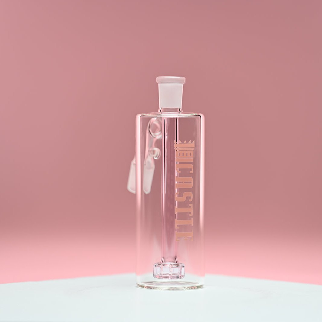 The image features a clear glass ash catcher with a light pink showerhead percolator inside. The brand name "CASTLE" is heat bonded vertically on the front of the ash catcher. The background is a soft, solid pink, which complements the gentle pink tone of the percolator. The ash catcher is placed on a white surface and is photographed from a front-facing angle, emphasizing the transparency of the glass and the delicate pink hue of the internal percolator.