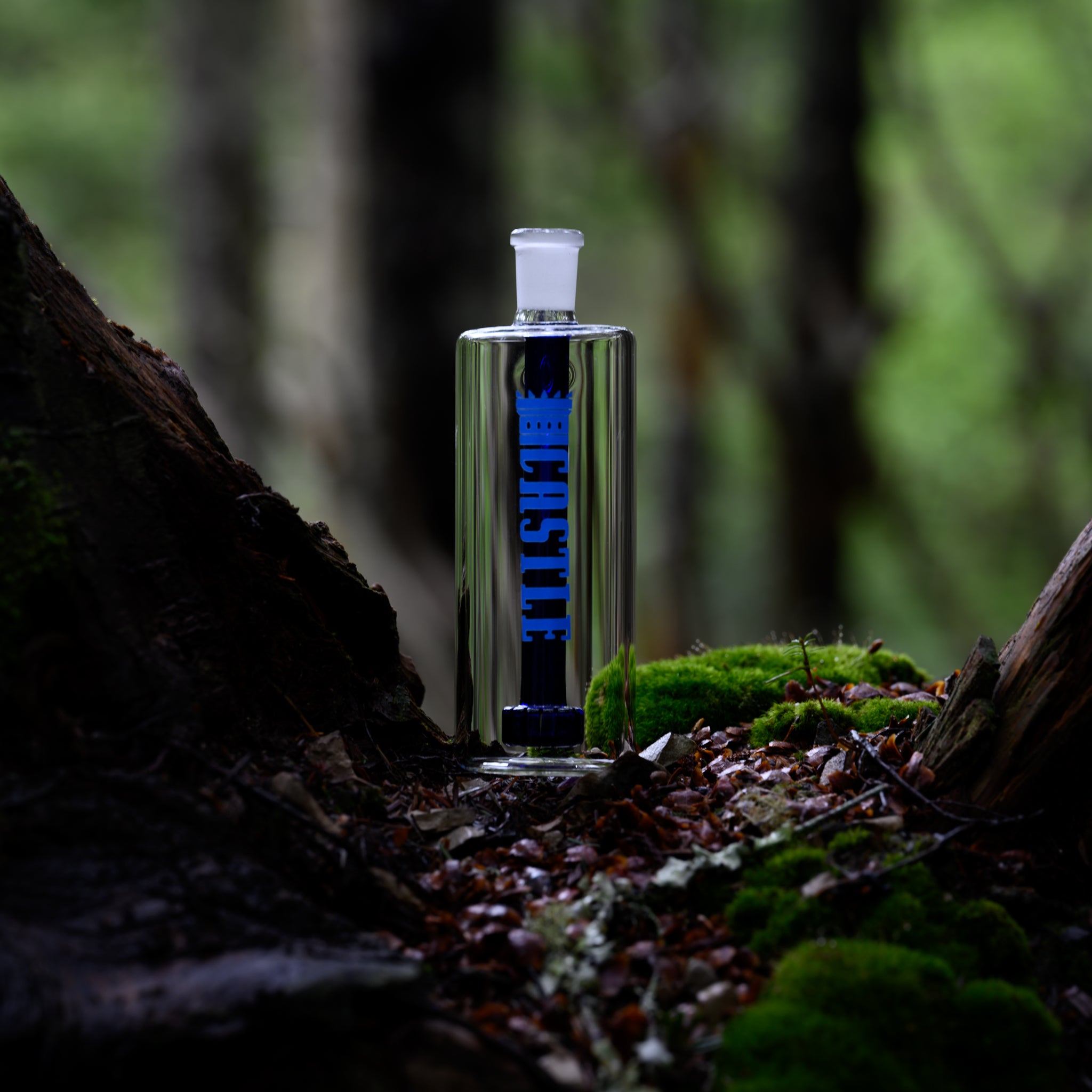 A clear glass ash catcher with blue accents and the brand name "Castle" in blue letters, positioned on a forest floor covered in fallen leaves and moss. The background features blurred trees, creating a tranquil, woodland atmosphere. The ash catcher stands upright, with its sleek design contrasting against the natural, earthy setting. The moss and leaves add texture and color to the scene, highlighting the ash catcher's clean lines and modern aesthetic.