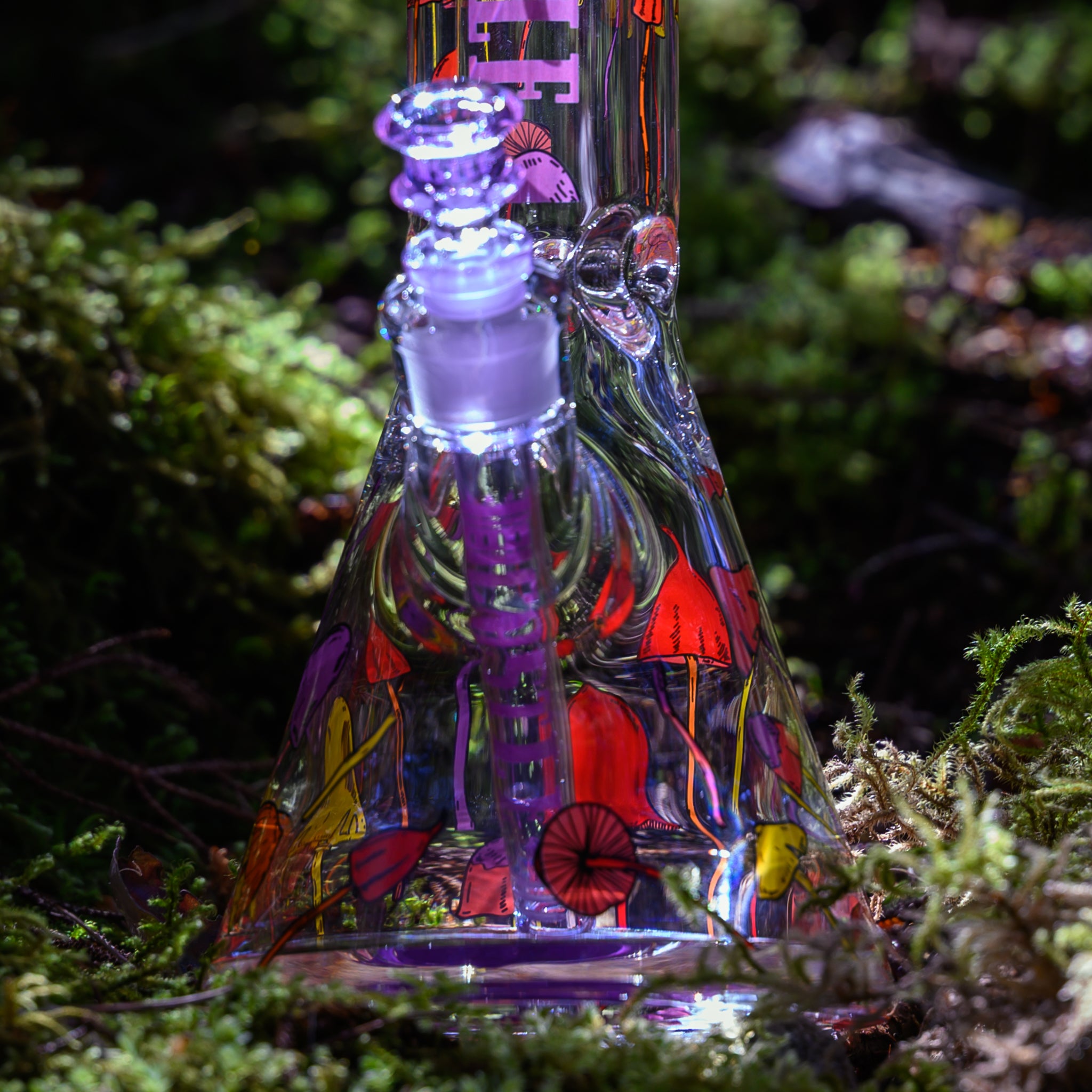 A close-up view of the base of a glass beaker bong with colorful mushroom designs. The downstem and bowl, colored purple, complement the whimsical artwork of mushrooms in various vibrant colors. The bong is placed on a forest floor, surrounded by lush green moss. The sunlight filtering through the trees highlights the intricate details of the glass and the vivid patterns, creating a magical and earthy atmosphere.