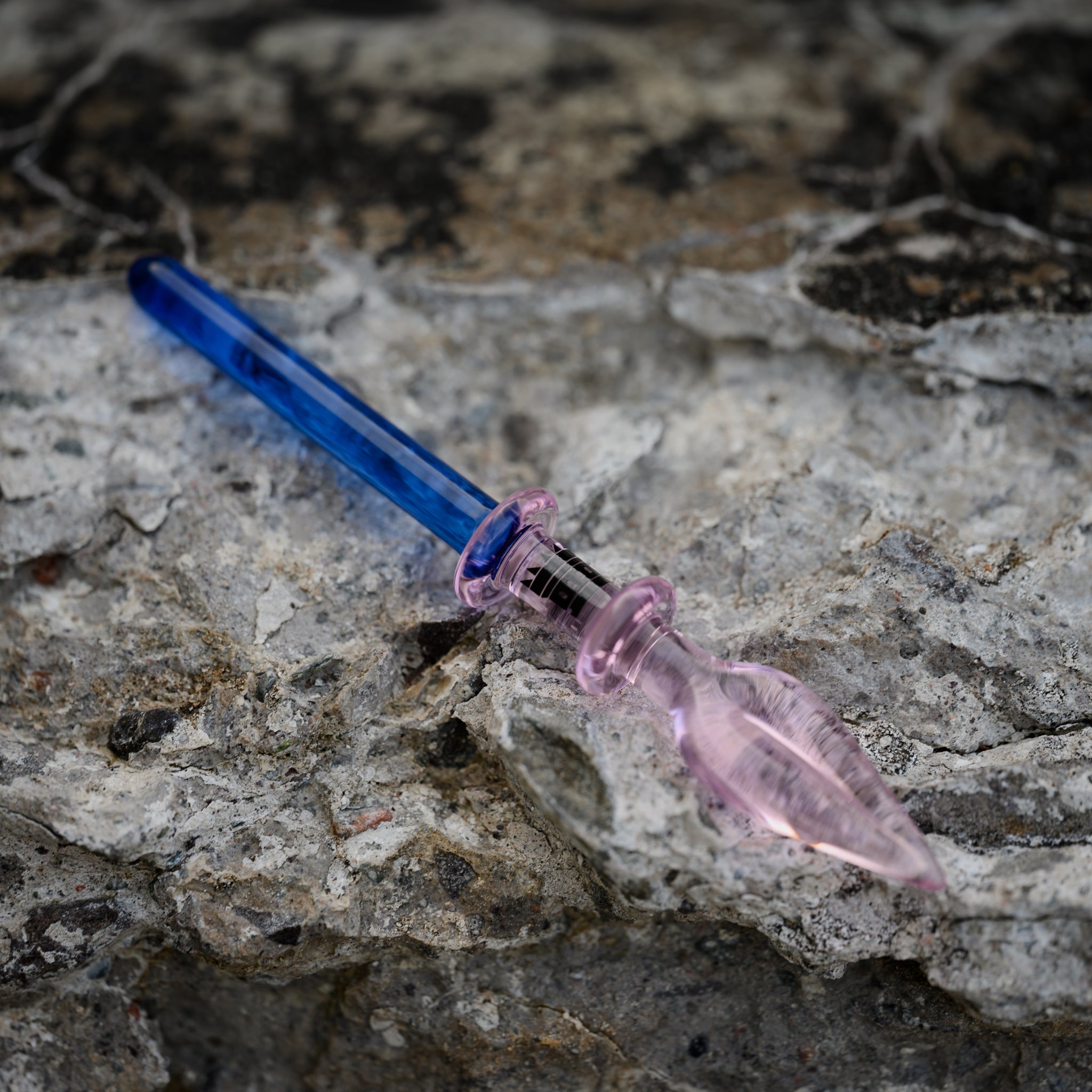 A blue and pink glass dab tool with a spade-shaped tip lies on a rough, textured concrete surface. The vibrant colors of the tool stand out against the neutral tones of the background, highlighting its intricate design and craftsmanship.