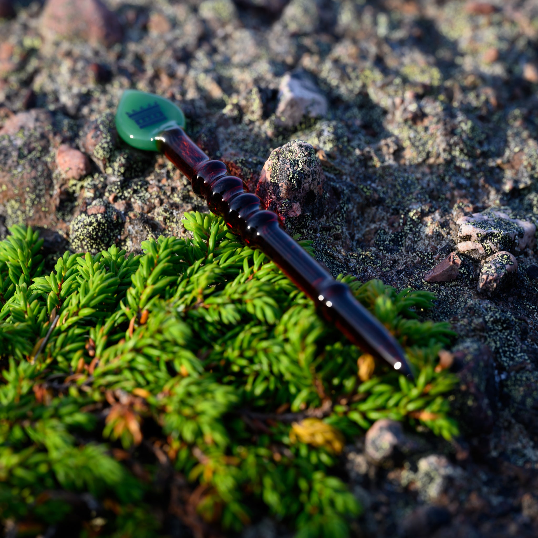 A dark, intricately designed glass dab tool with a spear-shaped tip lies on a rocky surface next to a vibrant patch of green moss. The tool's deep hues contrast strikingly with the lush greenery, emphasizing its detailed craftsmanship and unique appearance.