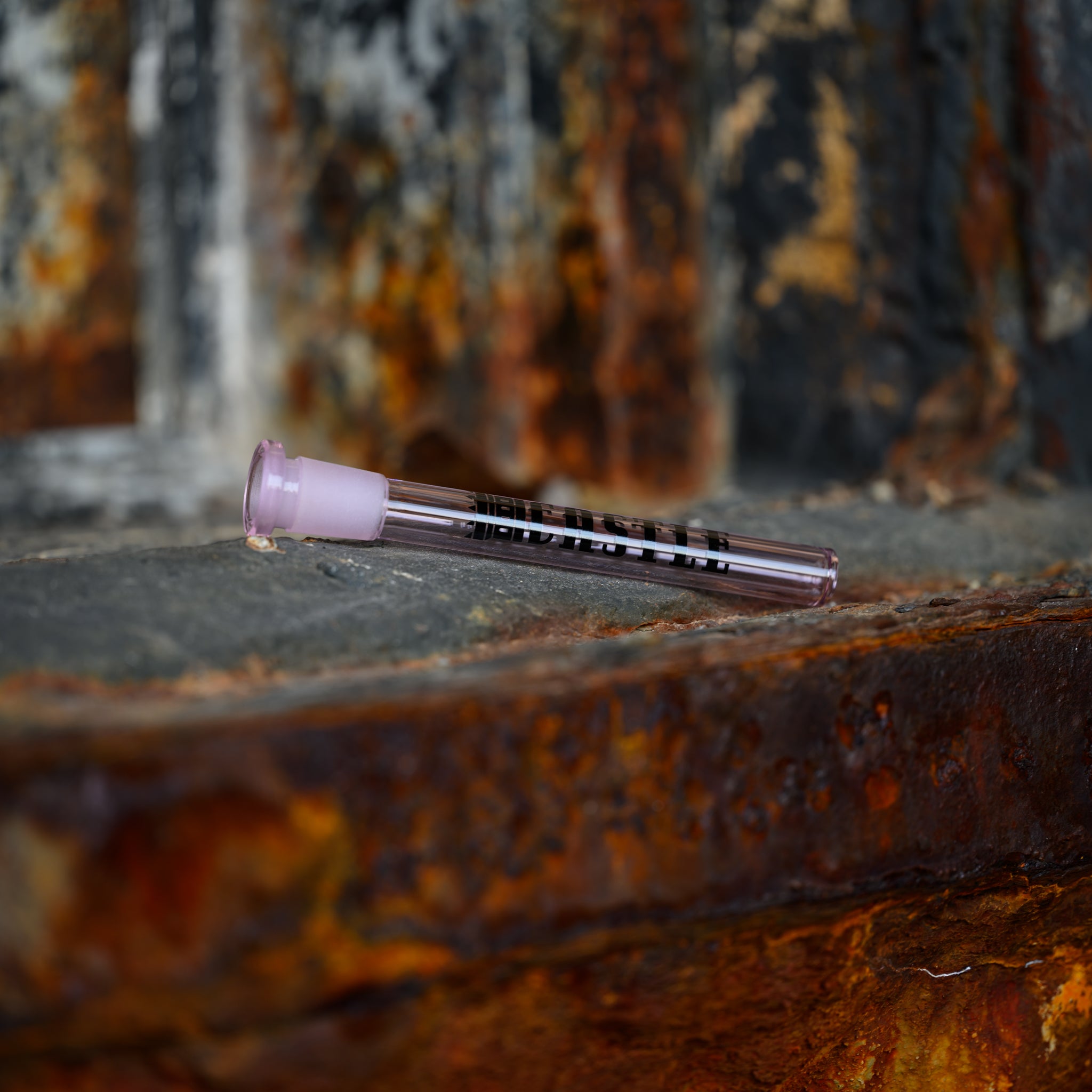 A pink glass downstem with the Castle Glassworks logo is placed horizontally on a rusted metal surface. The contrast between the delicate, transparent glass and the rugged, rusted metal background highlights the downstem's sleek and polished design. The scene is naturally lit, bringing out the intricate details and smooth finish of the downstem, while the rusted surface adds a rustic and industrial touch to the composition.