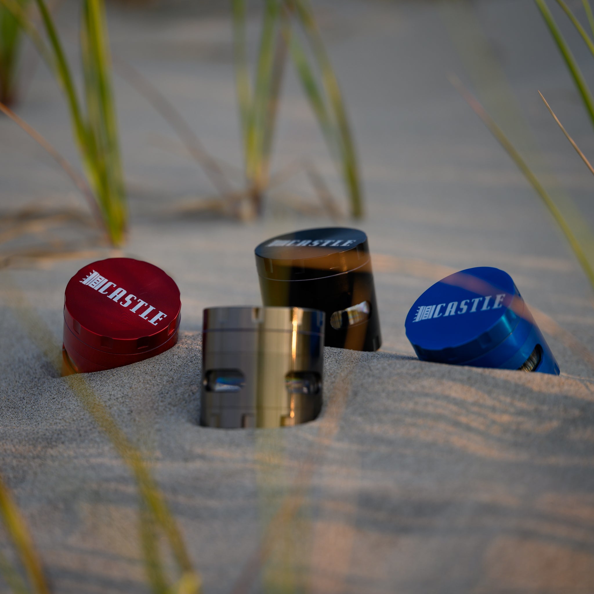 A set of four metal cannabis grinders, each with the "Castle" logo prominently displayed on the top, sitting partially buried in sandy ground. They are surrounded by tall blades of grass, which add a natural element to the setting. The soft evening sunlight casts gentle shadows, highlighting the grinders' sleek design and vibrant colors against the neutral sand backdrop.