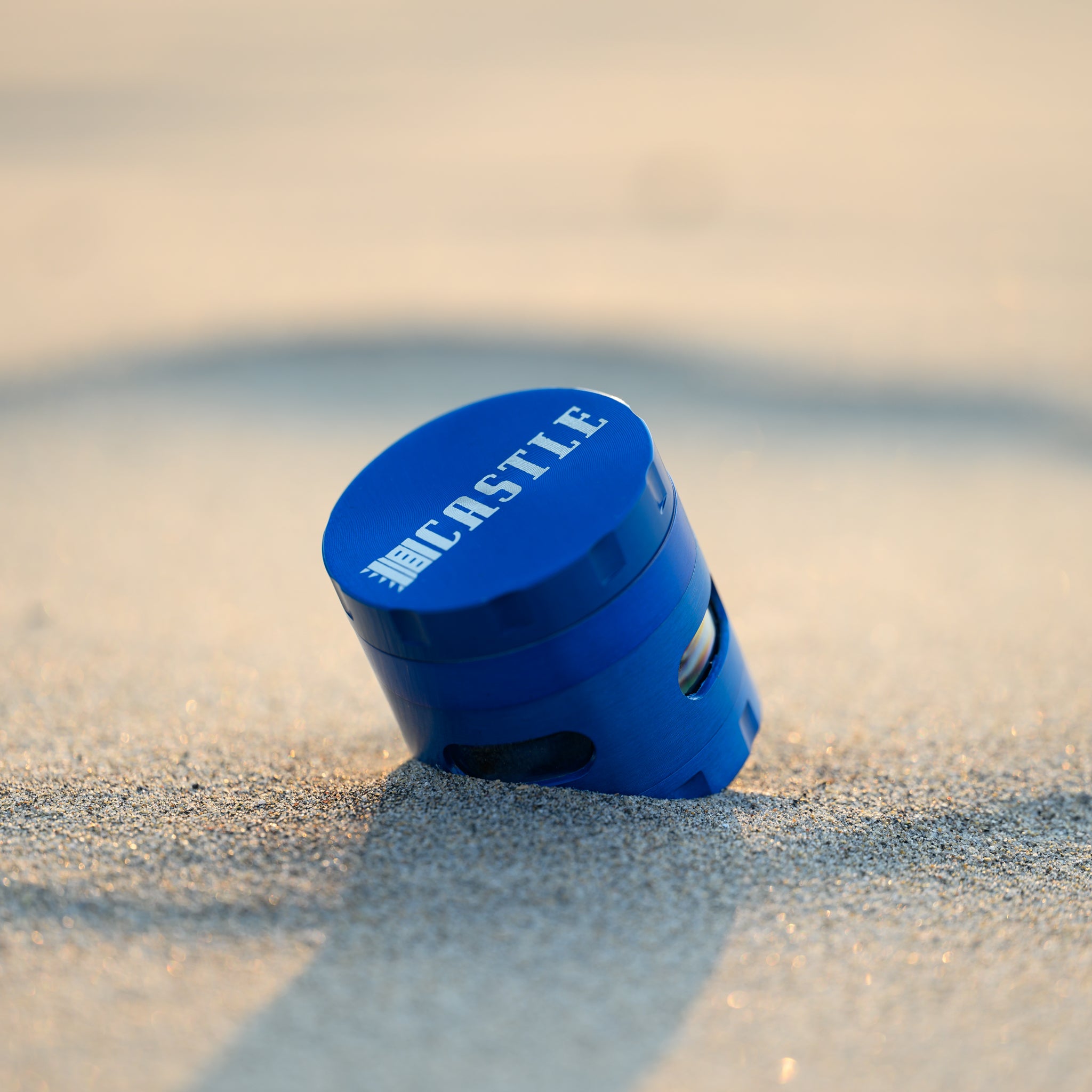 A close-up view of a blue metal cannabis grinder with the "Castle" logo on the top. The grinder is partially buried in the sand, tilted slightly to one side. The sunlight highlights the smooth finish and vibrant blue color, casting a soft shadow on the sand. The background is blurred, drawing focus to the detailed design of the grinder.