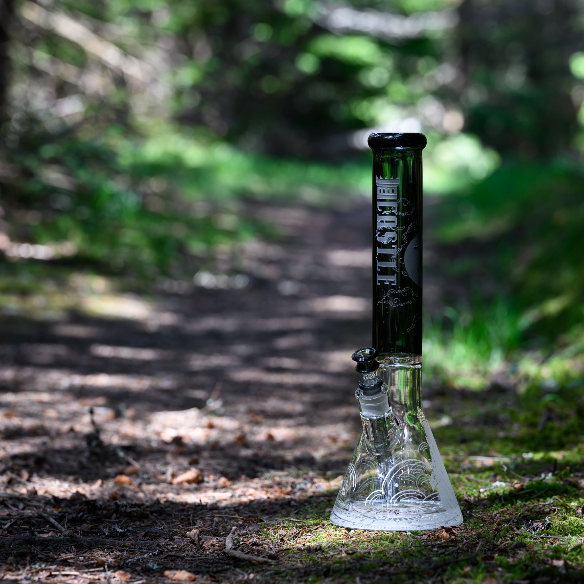 A glass beaker bong with a dark, smoky black neck and intricate designs featuring a wolf and wave patterns etched onto the base. The "Castle" logo is vertically displayed on the neck. The bong is placed on a forest path, with sunlight filtering through the trees and creating dappled shadows on the ground. The lush green foliage and natural surroundings provide a serene and earthy backdrop, highlighting the bong's detailed artwork and craftsmanship.