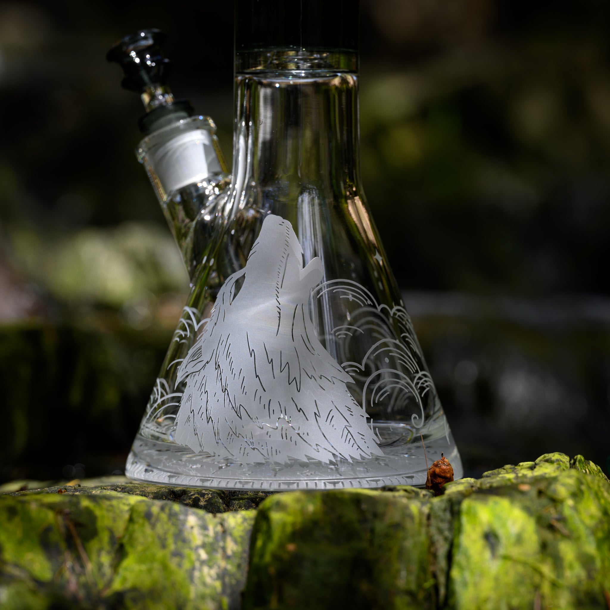 A close-up view of a glass beaker bong featuring an etched design of a howling wolf and wave patterns on the base. The bong is set against a backdrop of bright green moss-covered rocks, highlighting the detailed artwork and craftsmanship. The natural, earthy environment enhances the intricate design and creates a harmonious blend of nature and human-made art.